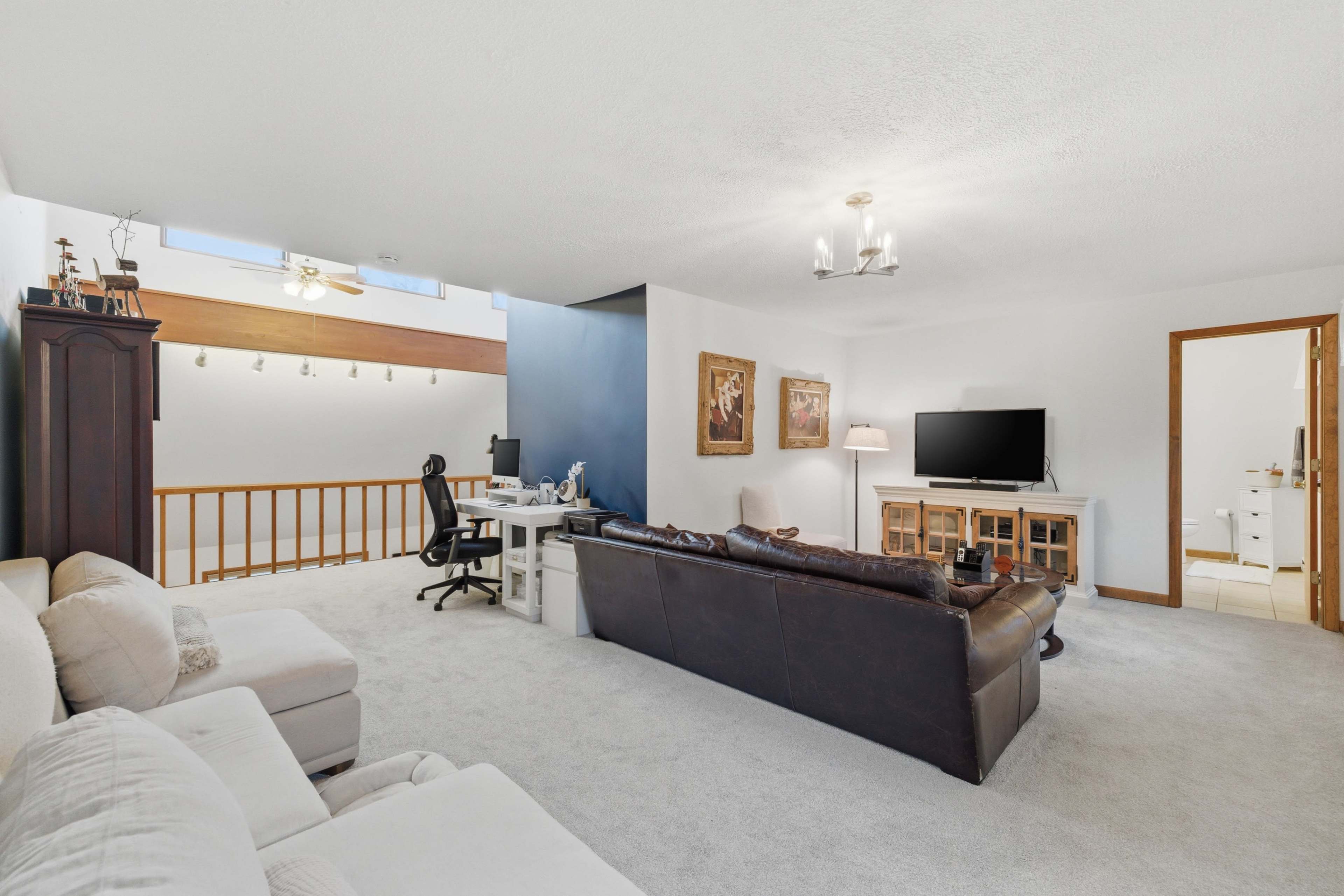 The image depicts a spacious living room with a dark leather sofa, a light-colored chair, a TV on a wooden stand, and a desk with a chair.