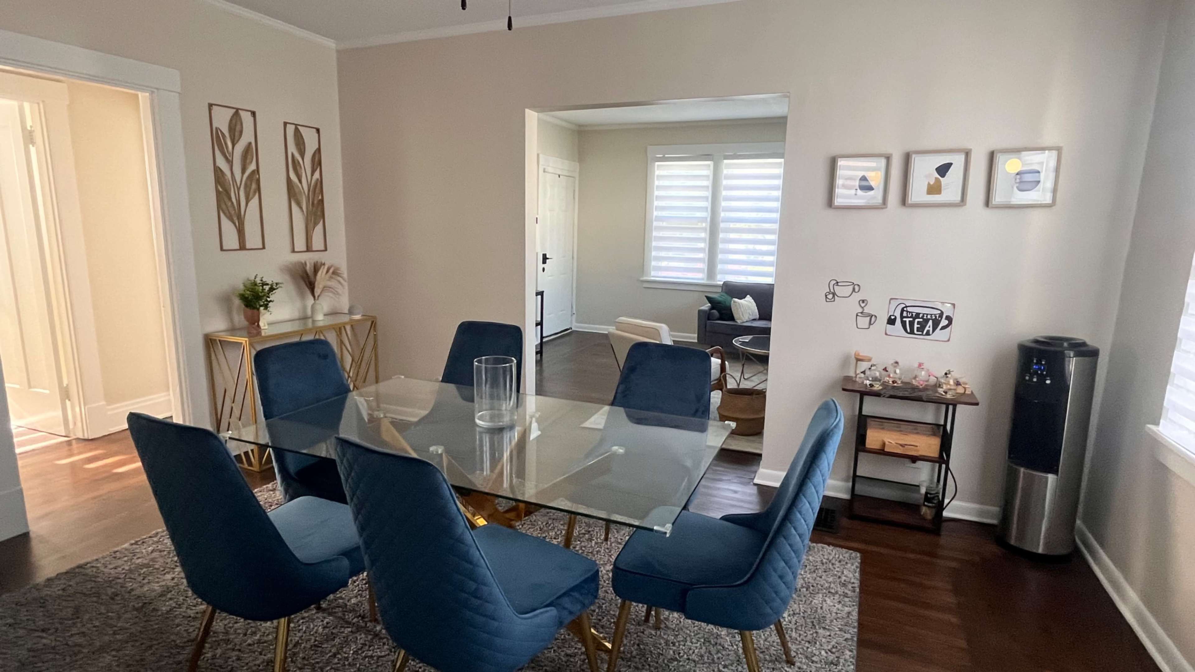 The image shows a modern dining area with a glass table surrounded by blue upholstered chairs, a small decorative plant, and framed artwork on the walls.