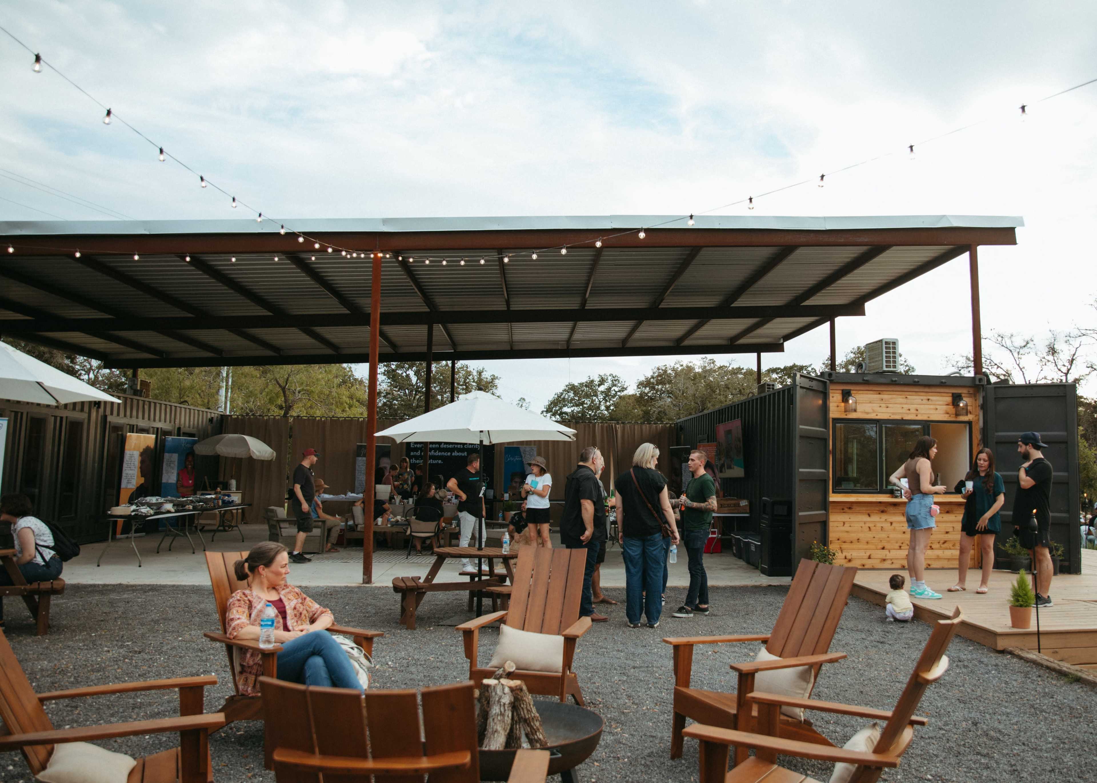 A gathering of people socializing in an outdoor space featuring wooden chairs, food trucks, and string lights overhead.