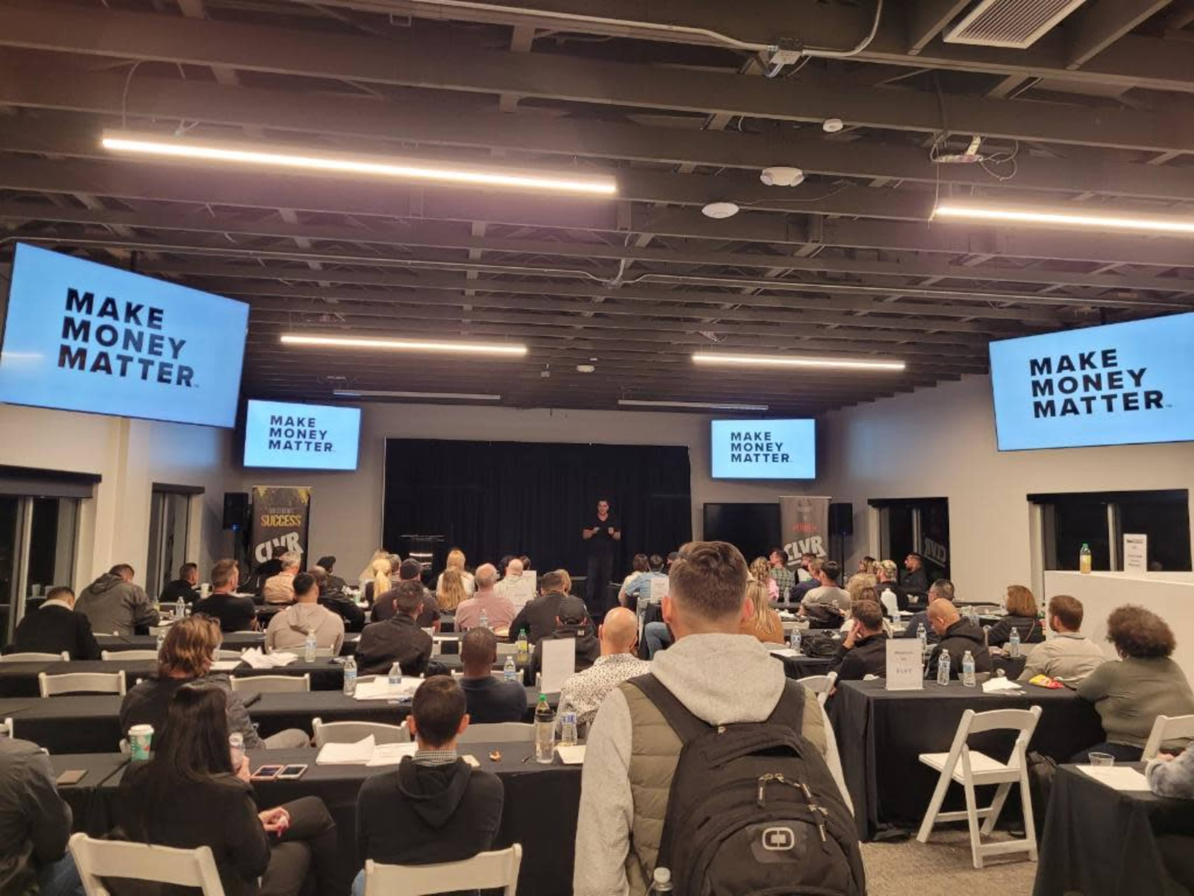 A speaker is addressing a large audience in a conference room with the phrase "MAKE MONEY MATTER" projected on screens.
