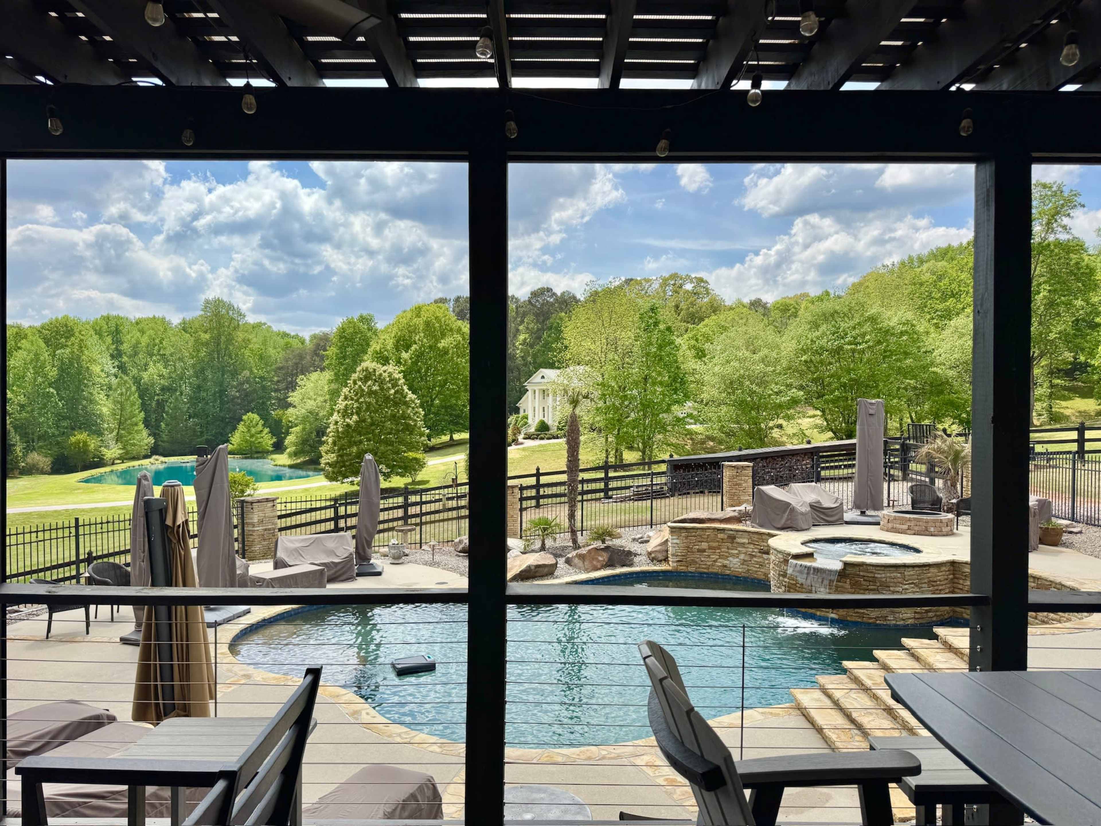 A view from a covered patio overlooks a swimming pool surrounded by lounge chairs, with lush greenery and a house in the background.
