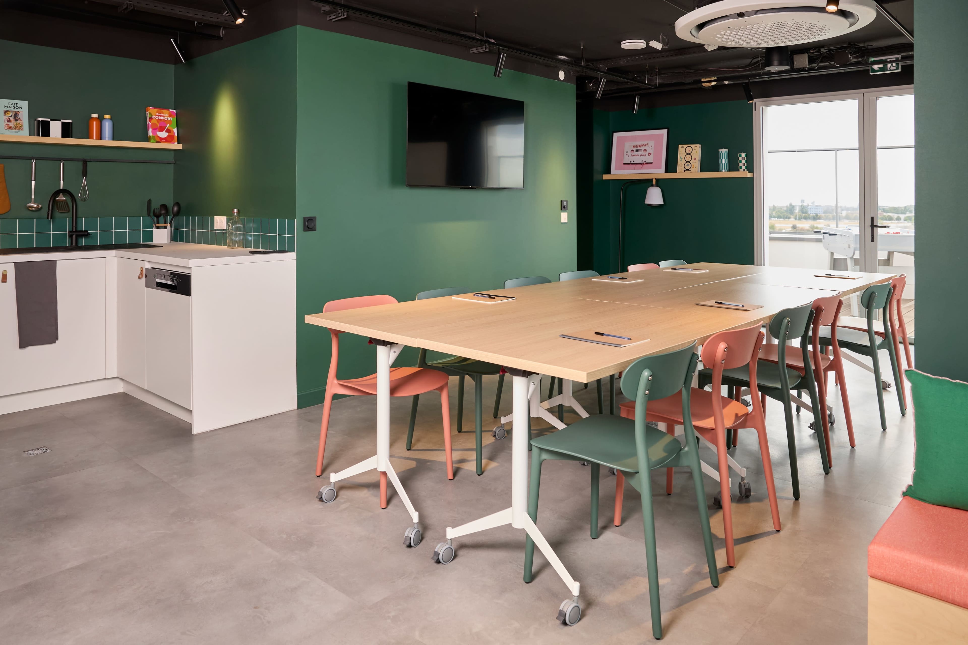 The image shows a modern meeting room with a long wooden table surrounded by colorful chairs, a kitchenette area, and large windows letting in natural light.