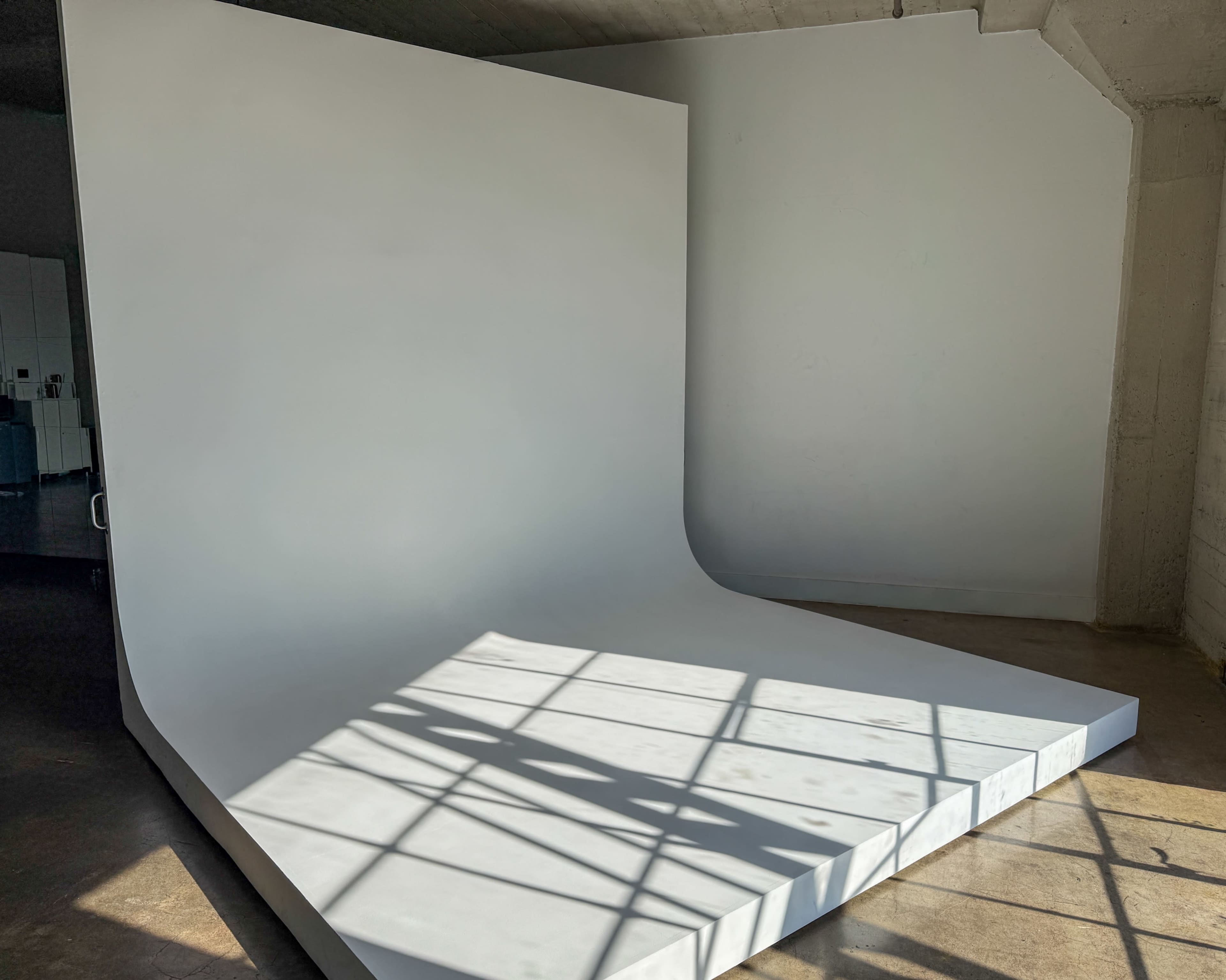 The image shows a spacious photography studio featuring a large white seamless backdrop with a sloped section and natural light casting shadows on the floor.