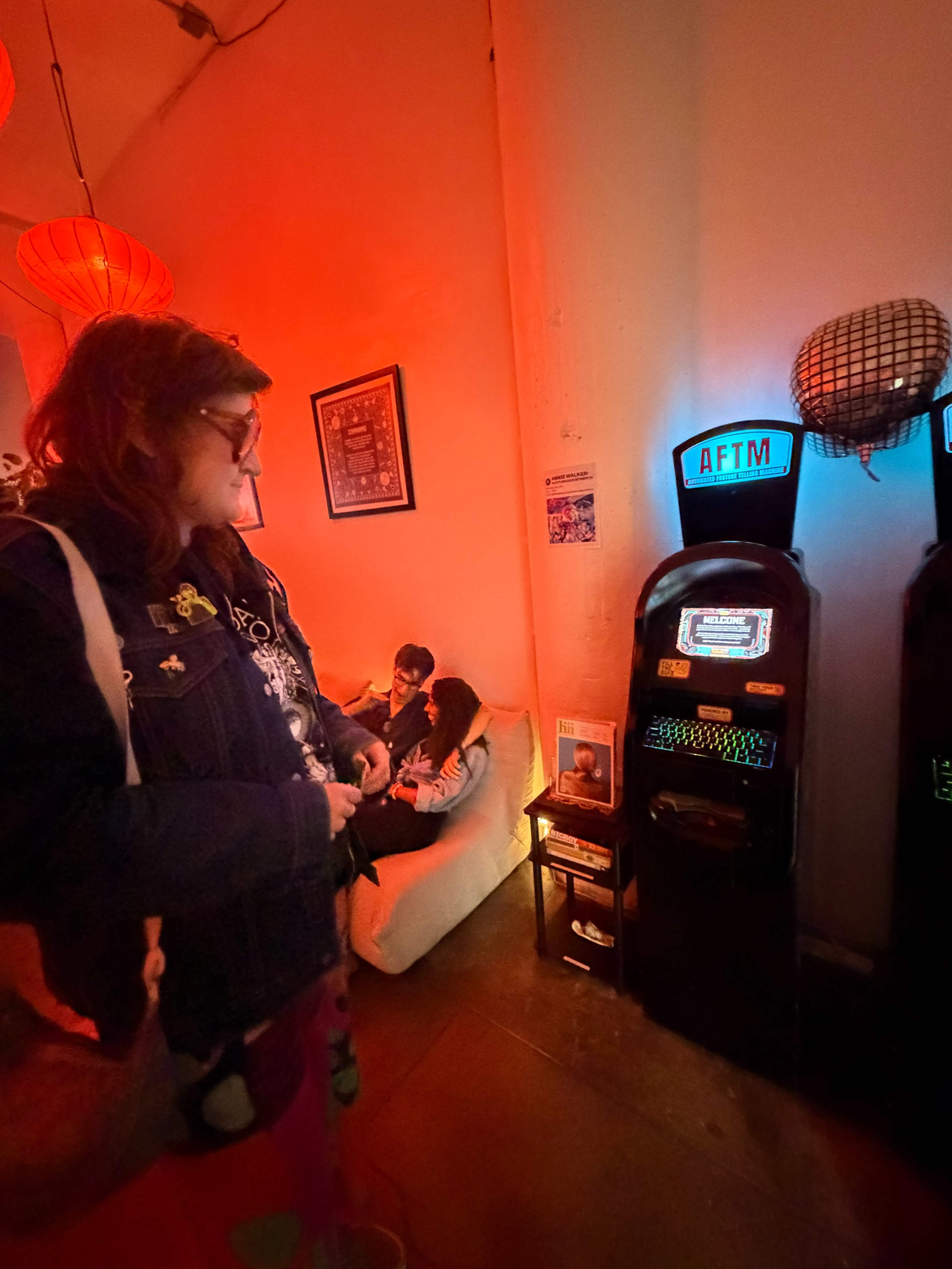 A woman stands next to a vintage arcade machine in a dimly lit room, while two people sit on a couch in the background.