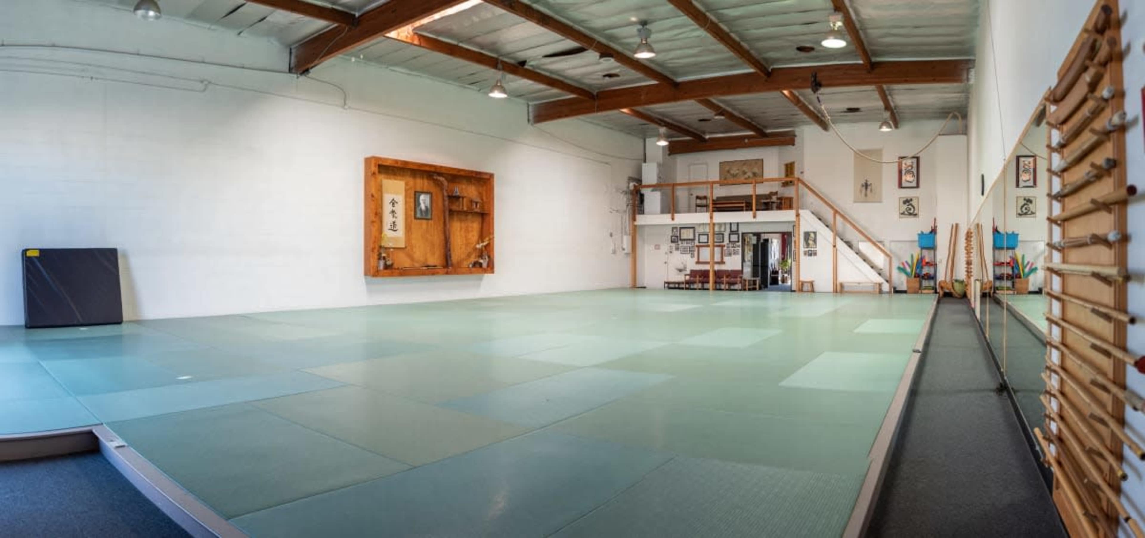 The image shows a spacious martial arts studio with tatami mats, wooden beams, and various wall decorations.