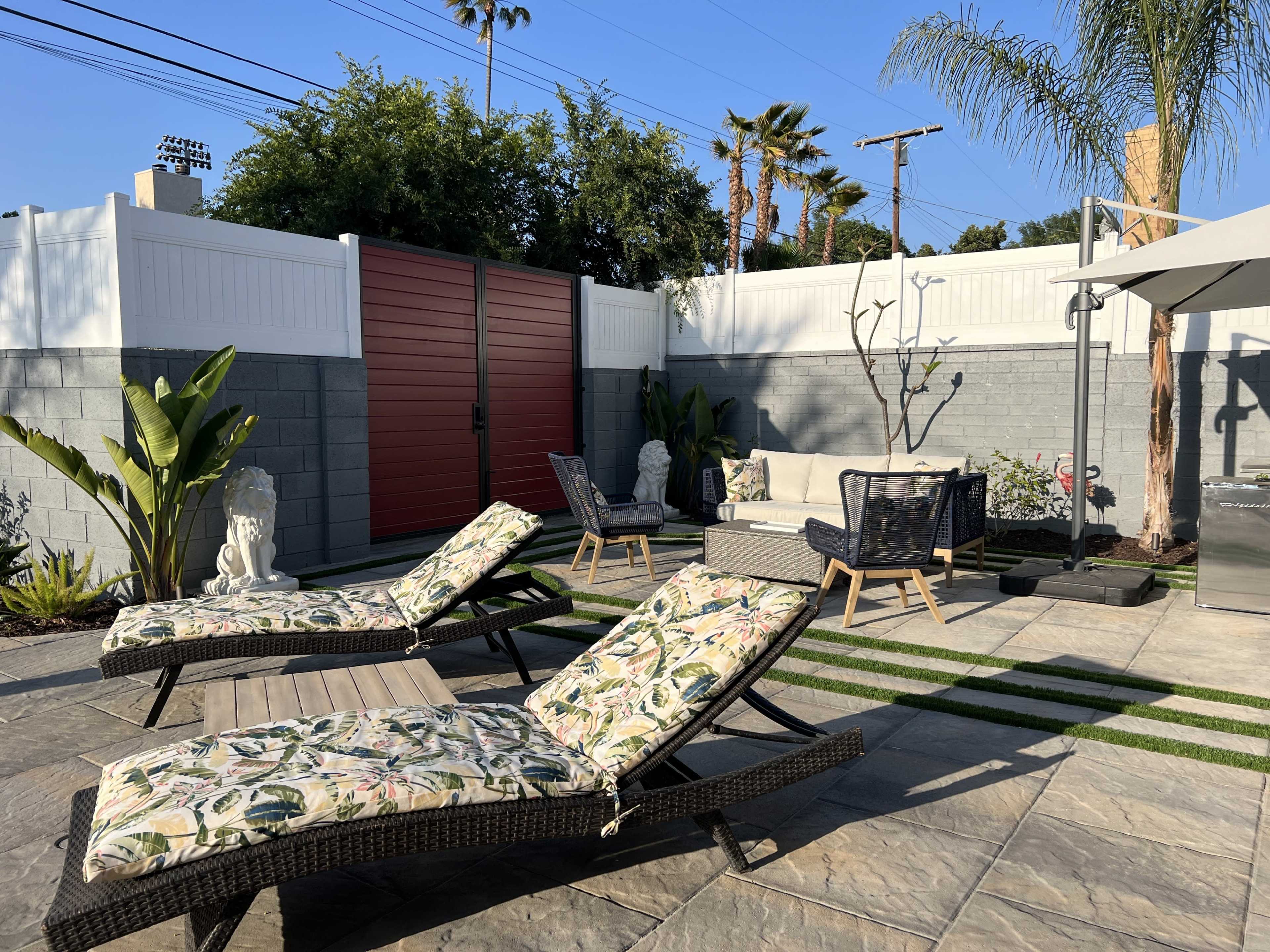 A landscaped backyard features lounge chairs, a seating area with a sofa, and decorative plants against a backdrop of a privacy fence and palm trees.