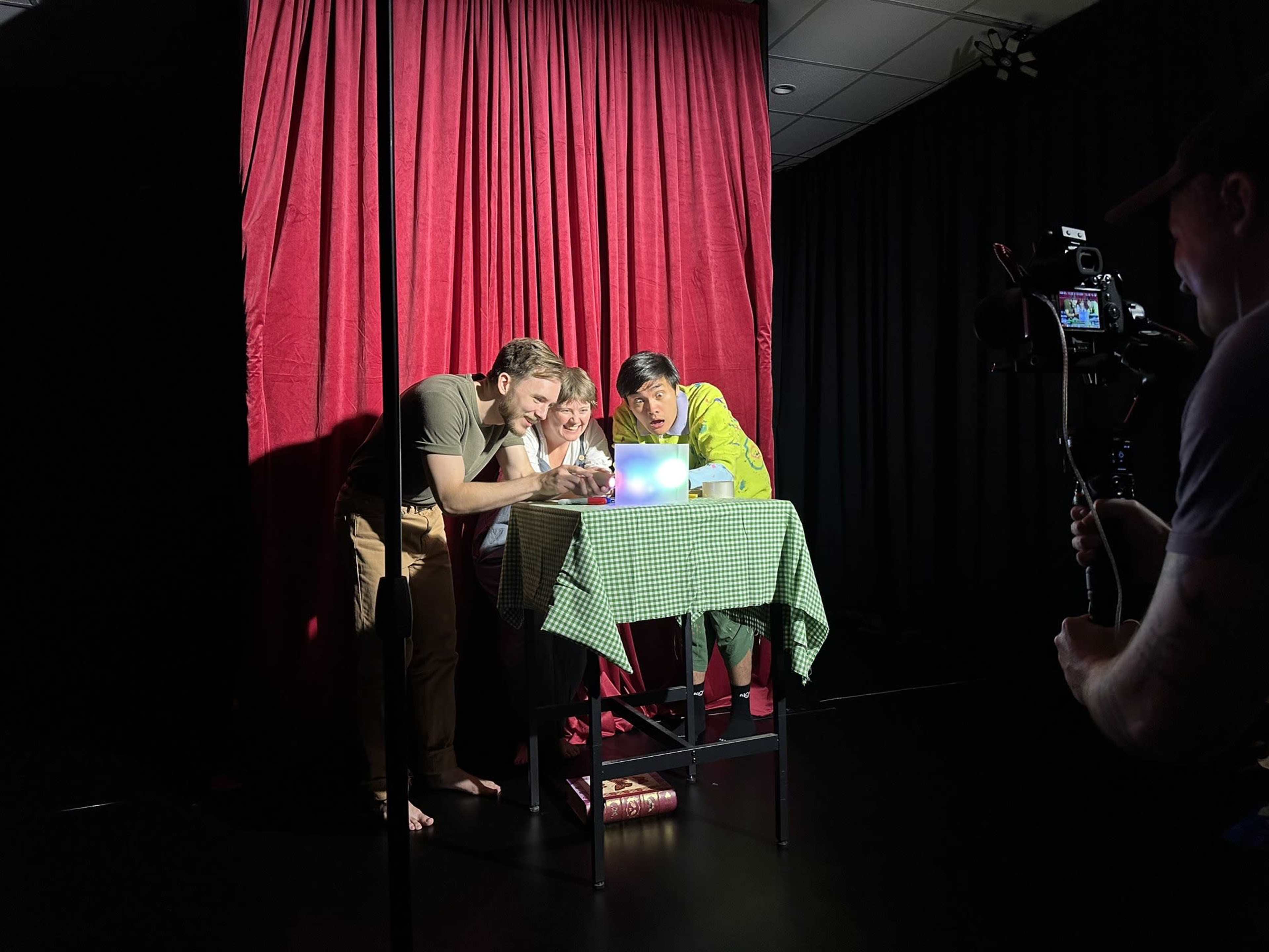 Three people are gathered around a table with a colorful light source, while a person behind a camera captures the scene against a backdrop of red curtains.