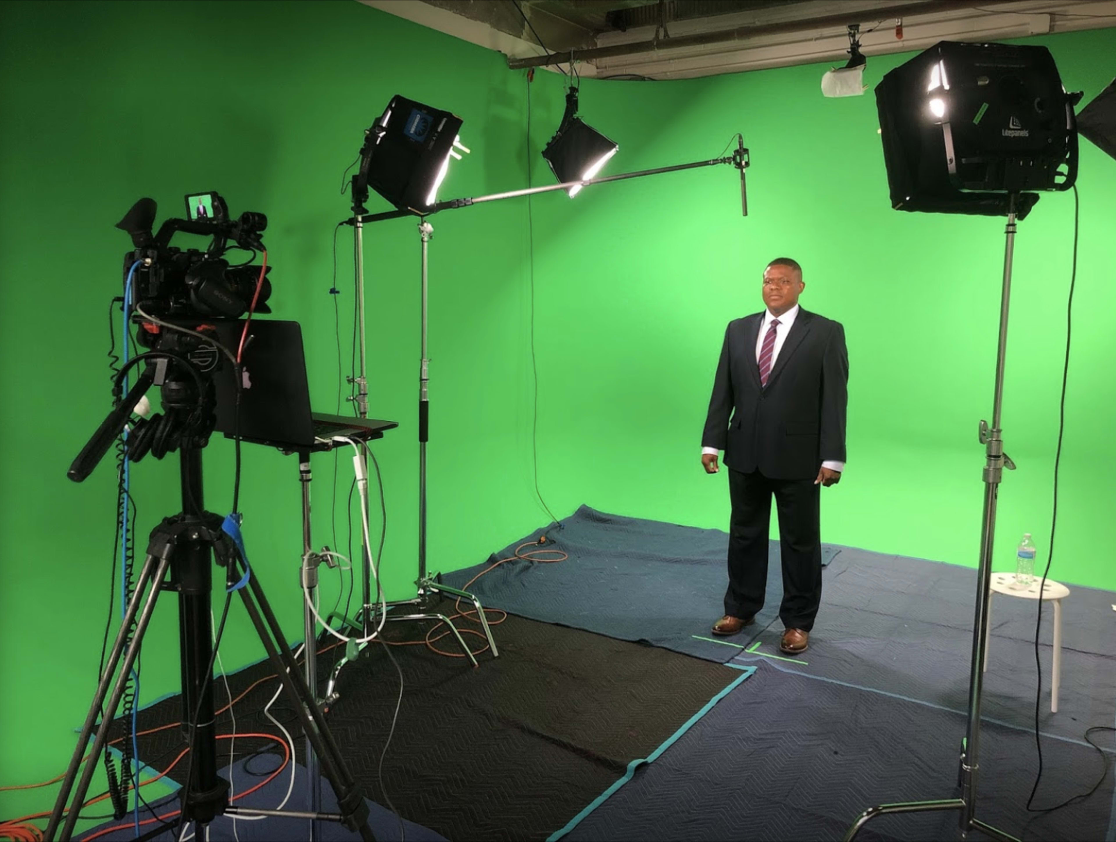 A man in a suit stands on a carpeted floor in front of a green screen, with cameras and lights set up nearby.