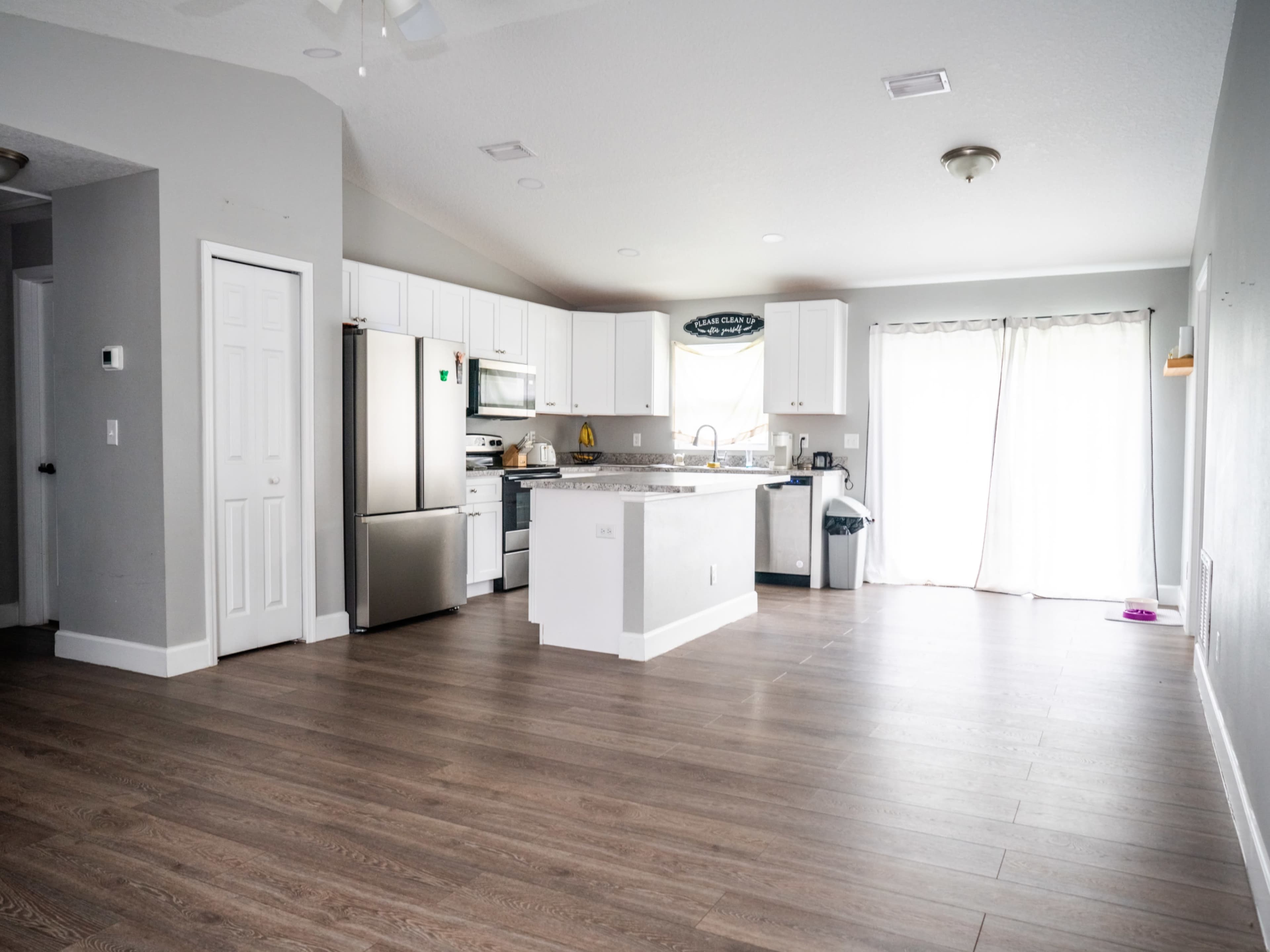 The image shows a spacious, modern kitchen with stainless steel appliances and light-colored cabinetry, featuring an open layout and a large window letting in natural light.