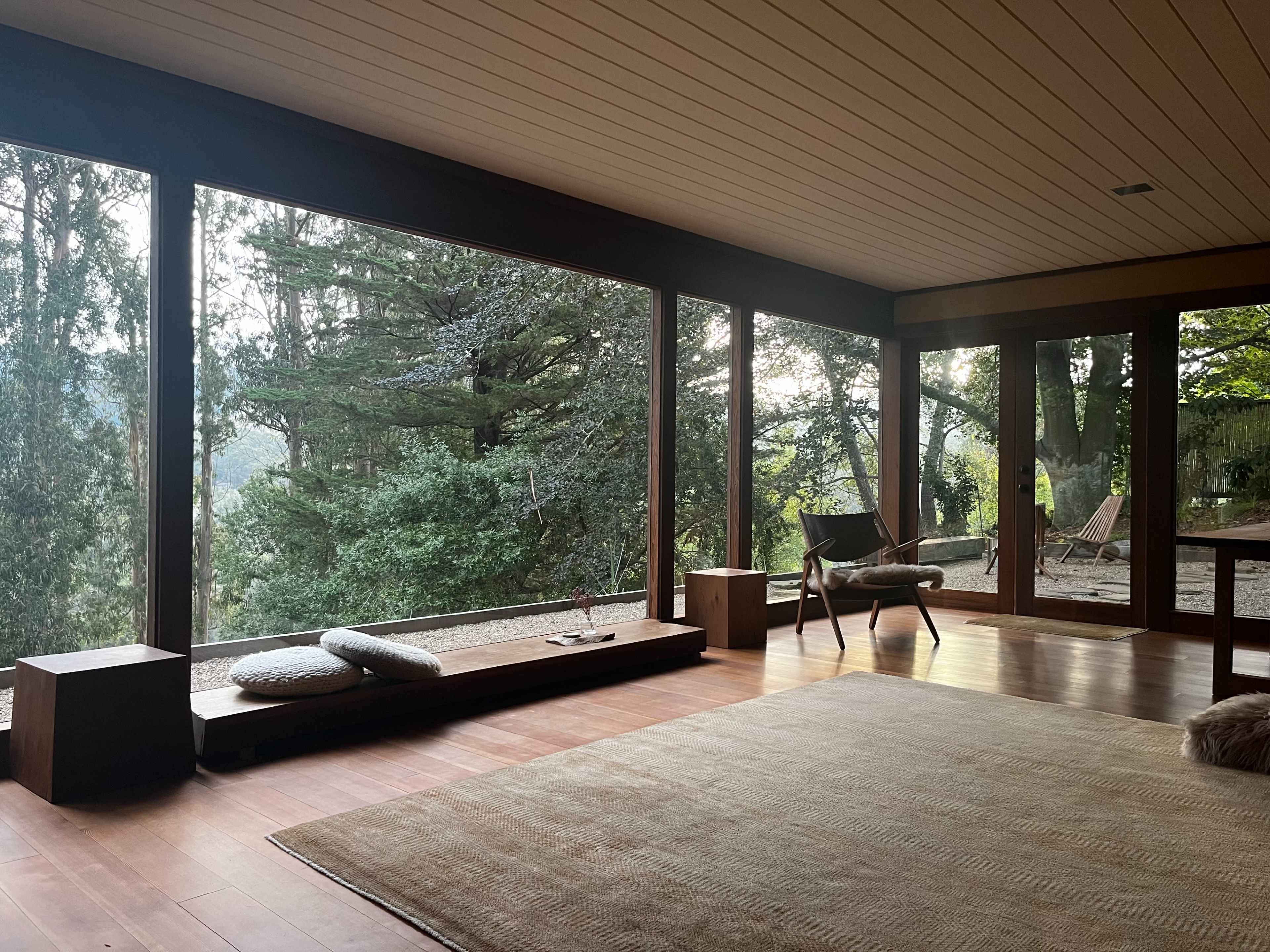 The interior features a spacious room with large windows overlooking a wooded landscape, wooden flooring, and minimal furniture.