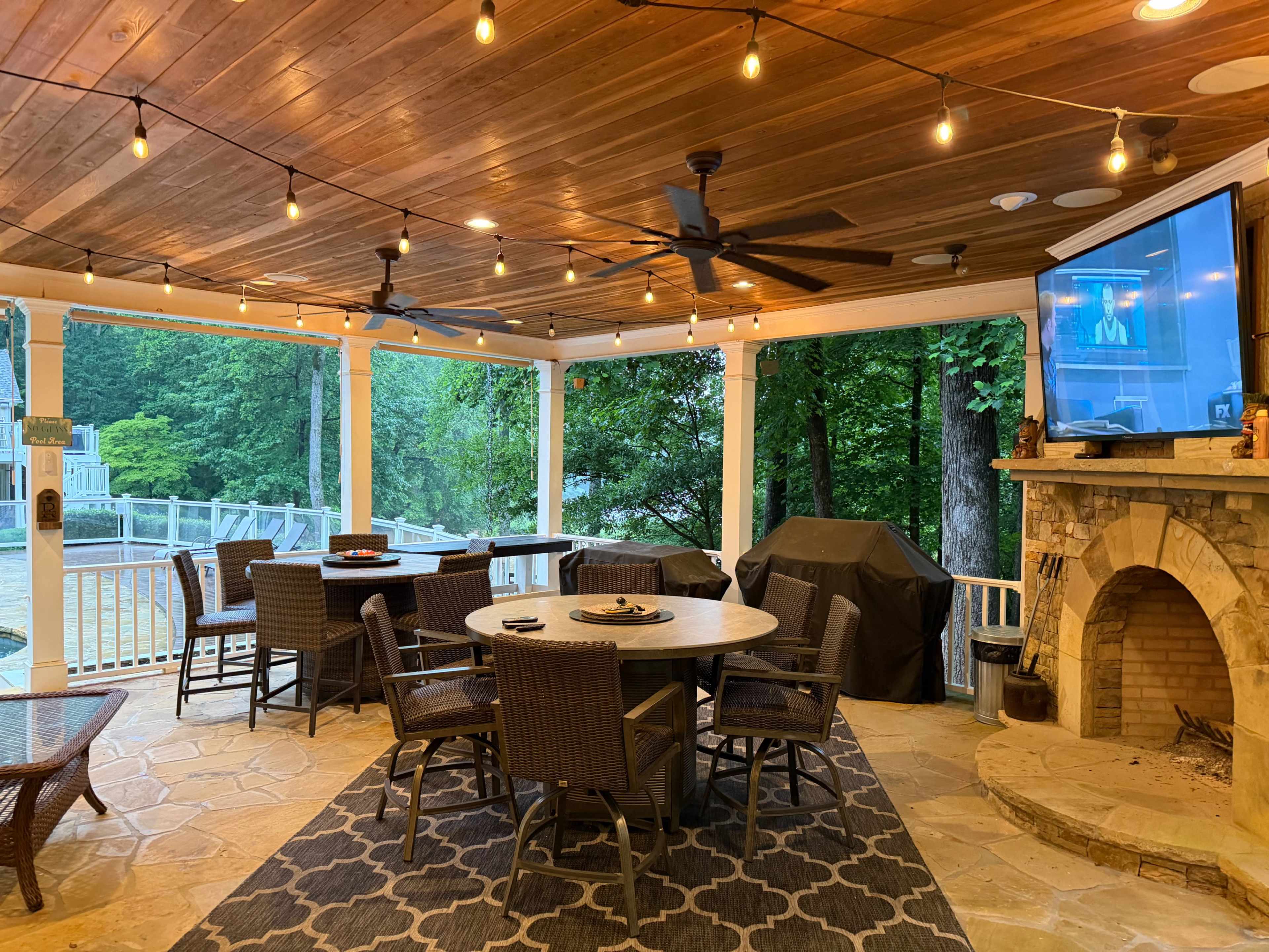 The image shows a covered patio area with a large table and several chairs, outdoor lighting, a grill, and a television mounted on the wall.