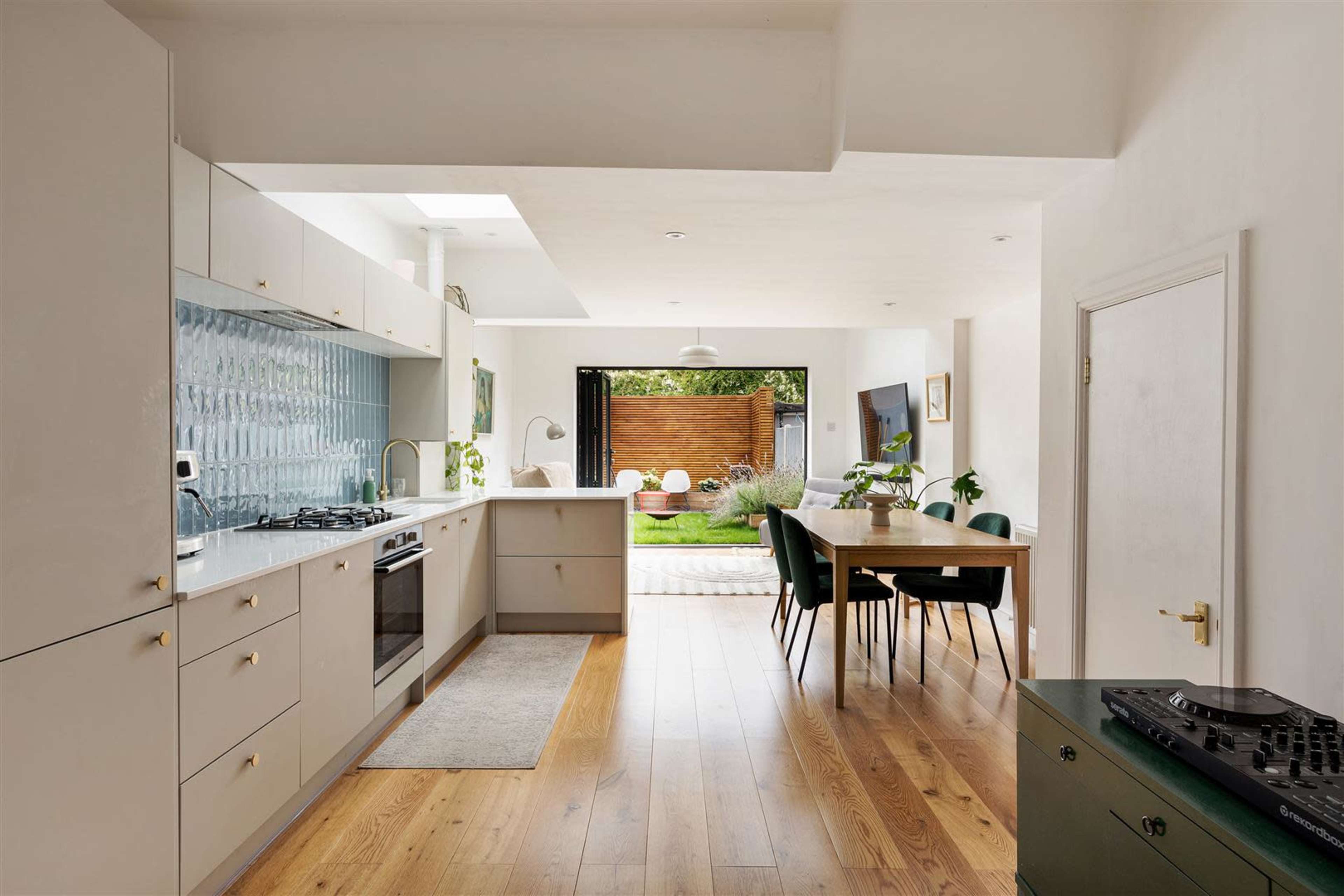 The image shows a modern kitchen with light cabinetry and an open layout leading into a dining area, featuring a table and chairs, with large windows overlooking a garden.