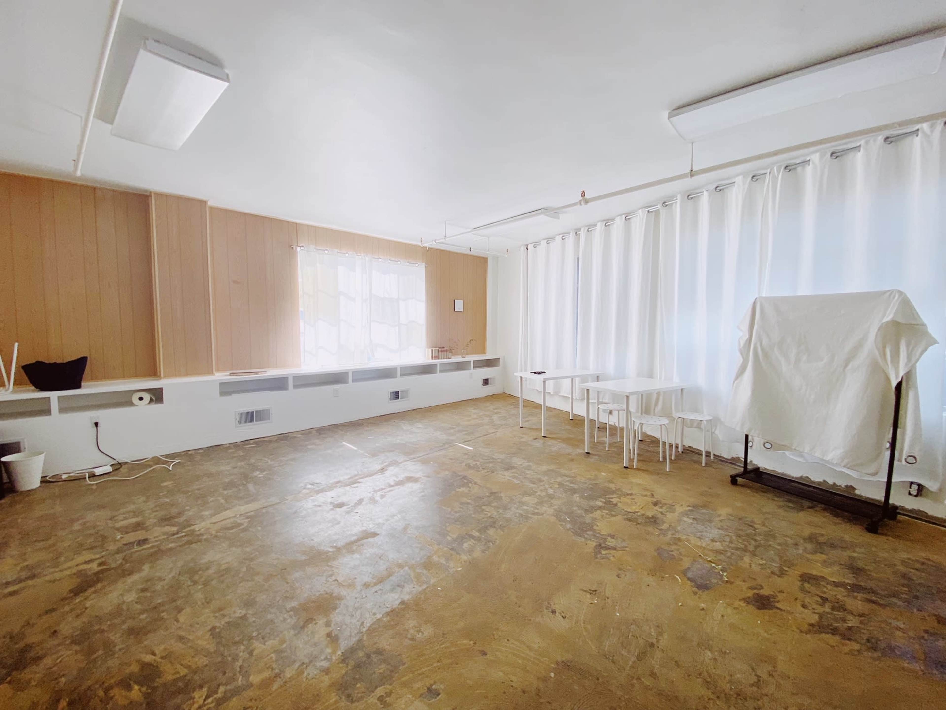 A spacious room with a polished concrete floor, large windows covered by sheer white curtains, and minimal furniture including a table and several chairs.