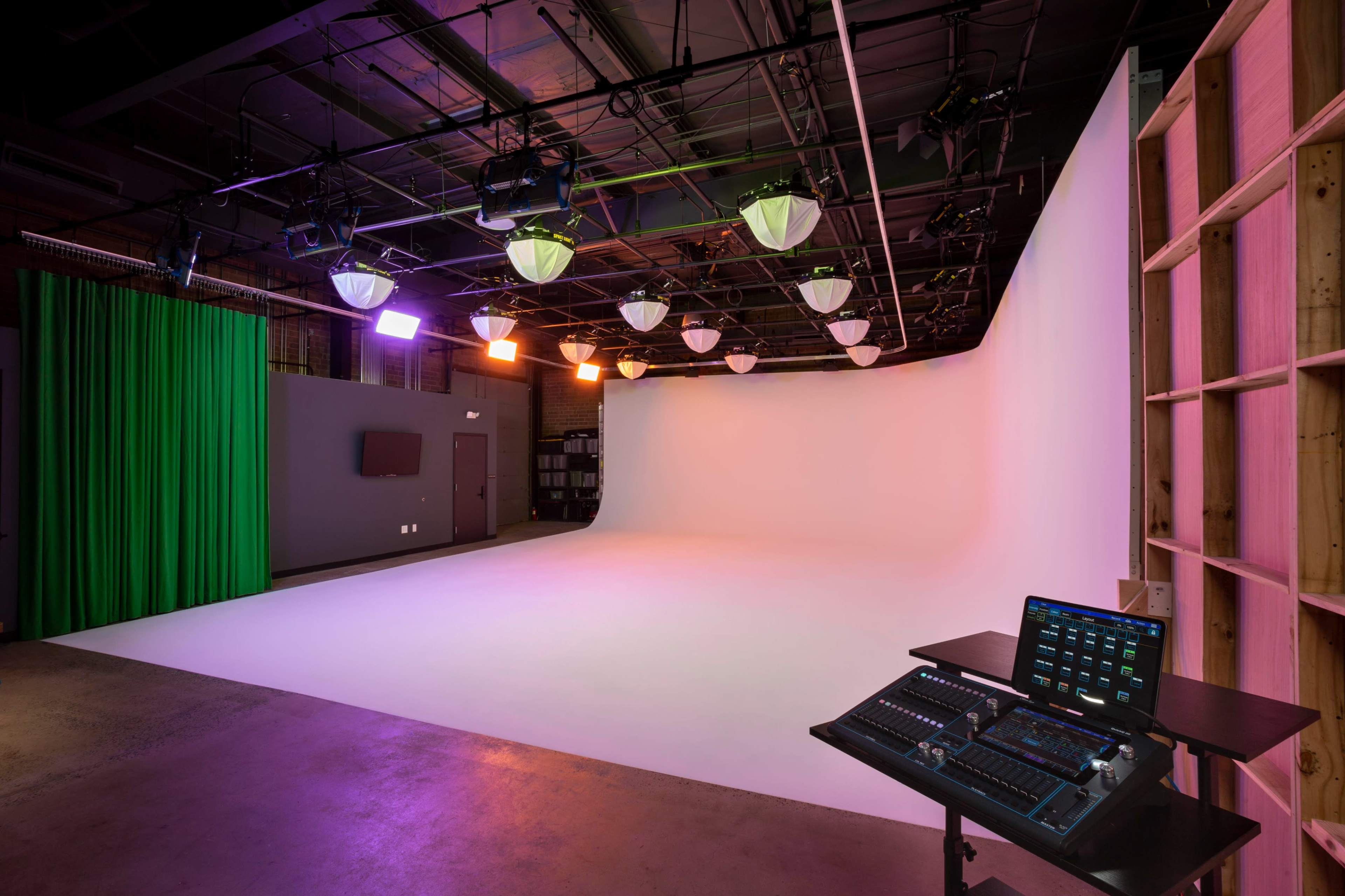 The image shows a modern studio with a white backdrop, green curtain, and lighting fixtures, along with a control panel on the side.