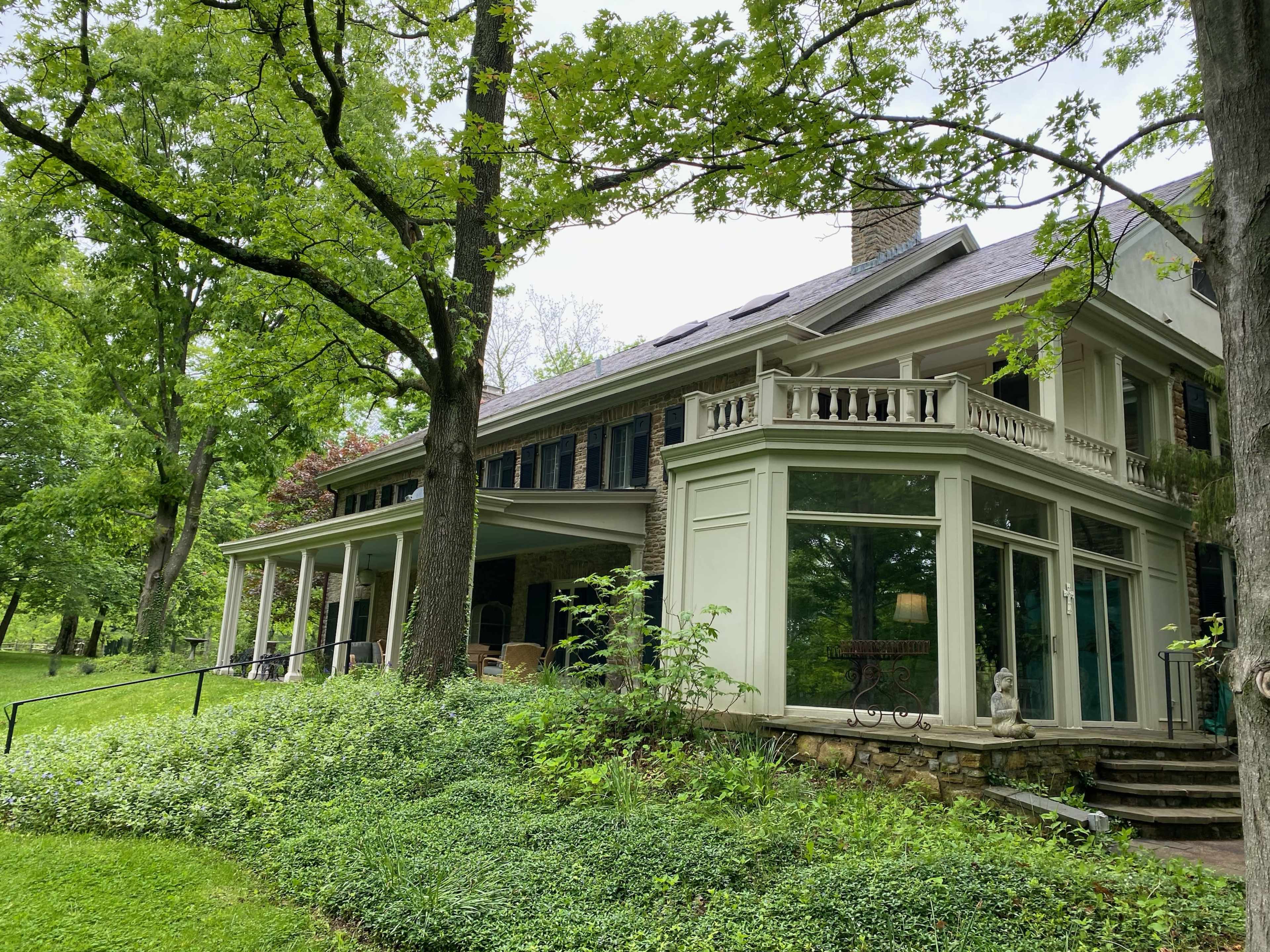A large, two-story house surrounded by green trees and shrubs, featuring a porch and multiple windows.