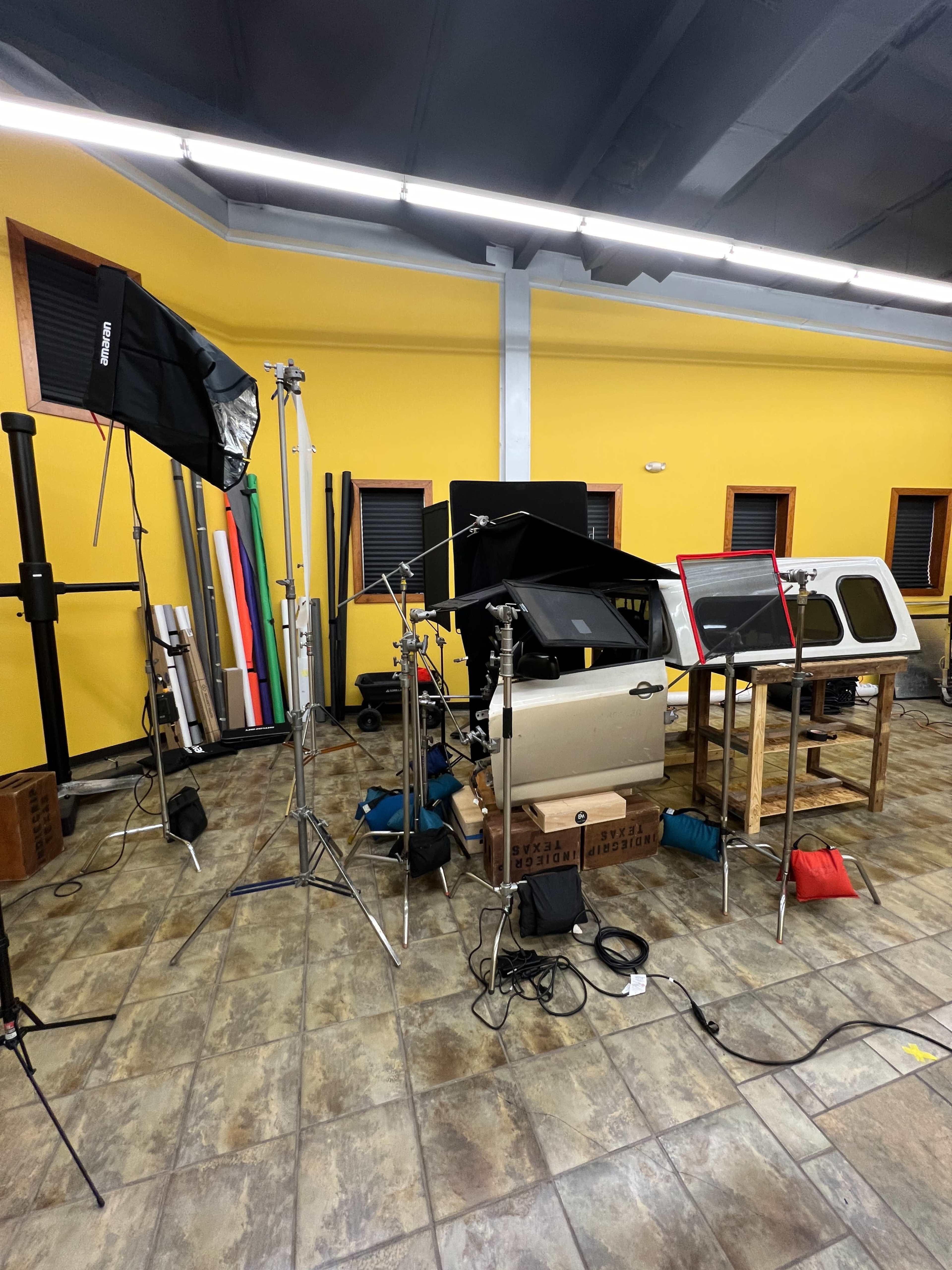 The image shows a studio setup with three light stands, a backdrop of colored materials, and various equipment arranged around a table featuring vehicle parts.