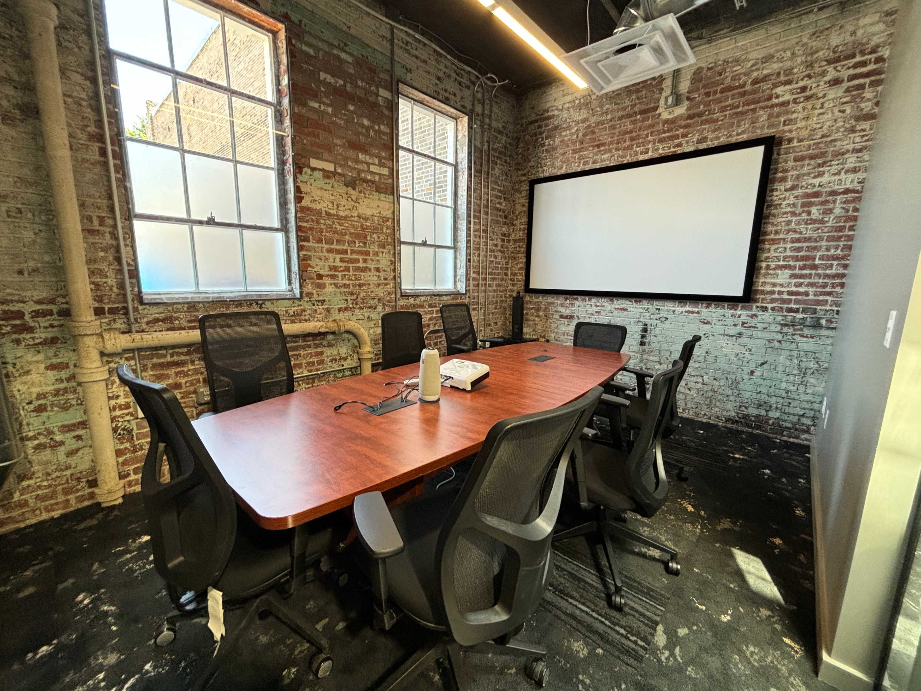 A conference room features a long wooden table surrounded by black chairs, with exposed brick walls and a large screen on one side.