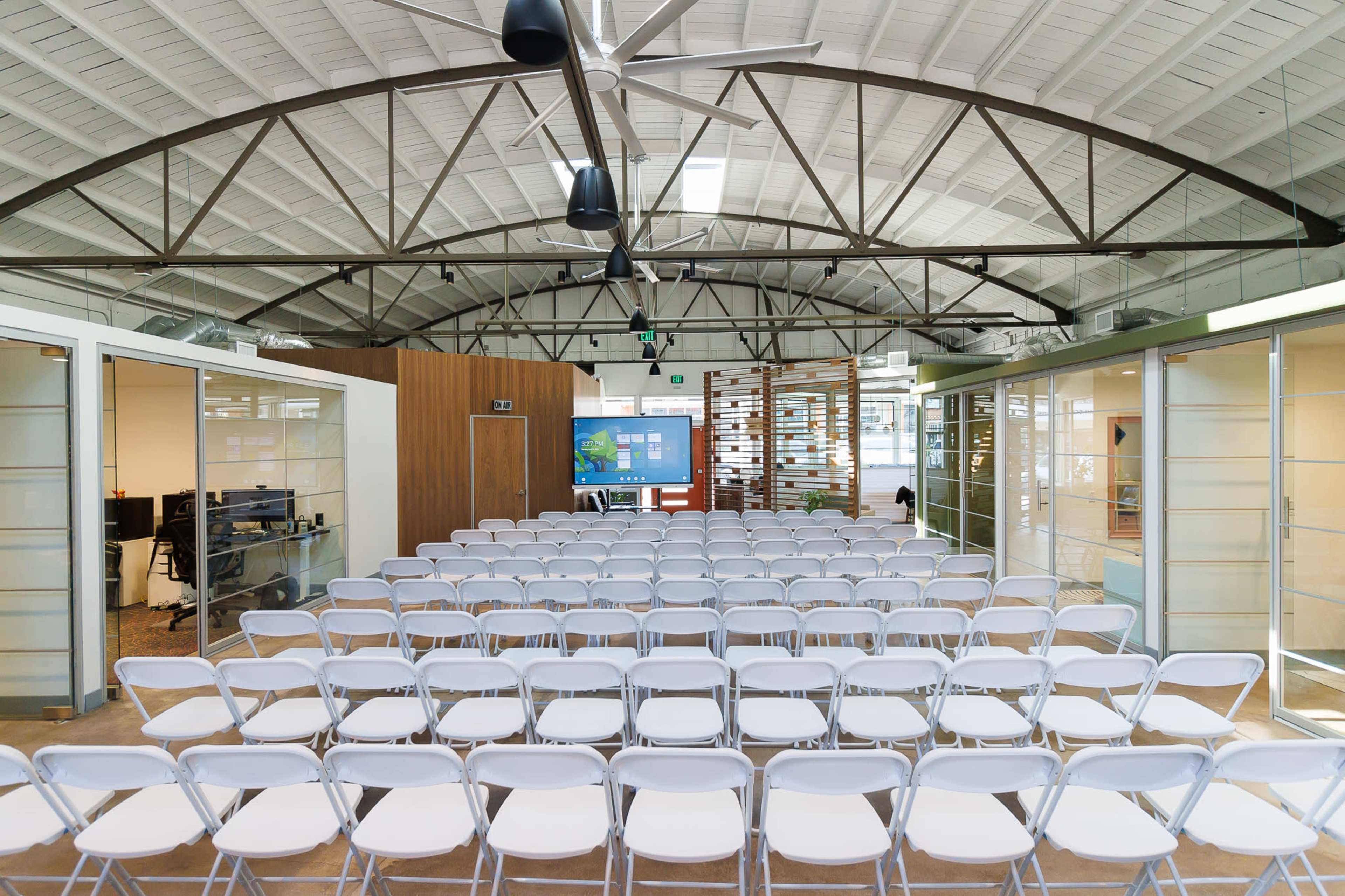 A spacious room with white folding chairs arranged in rows facing a presentation screen, flanked by glass-walled office spaces.