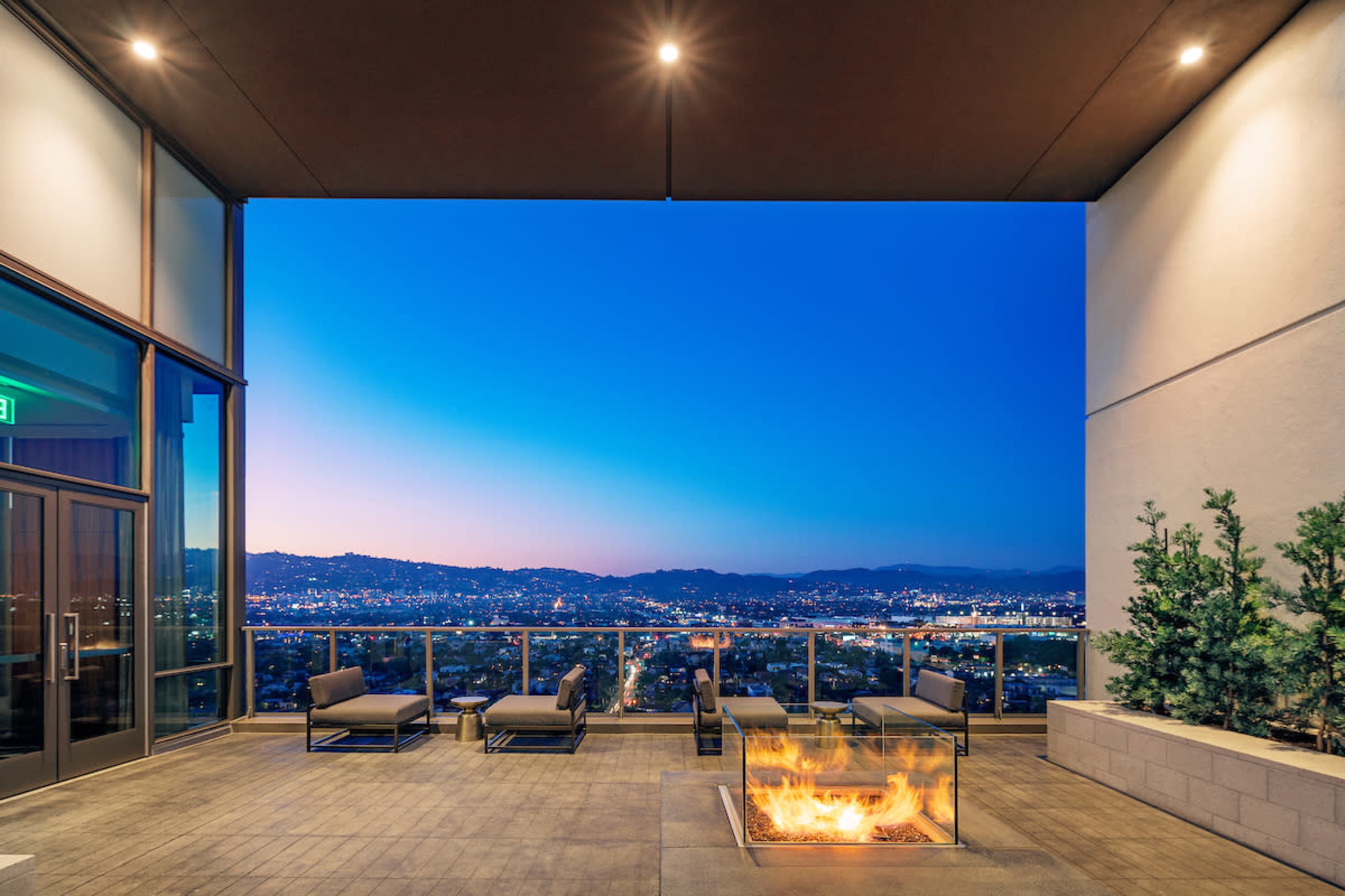 The image shows a modern balcony with seating and a fire pit, overlooking a cityscape at twilight.