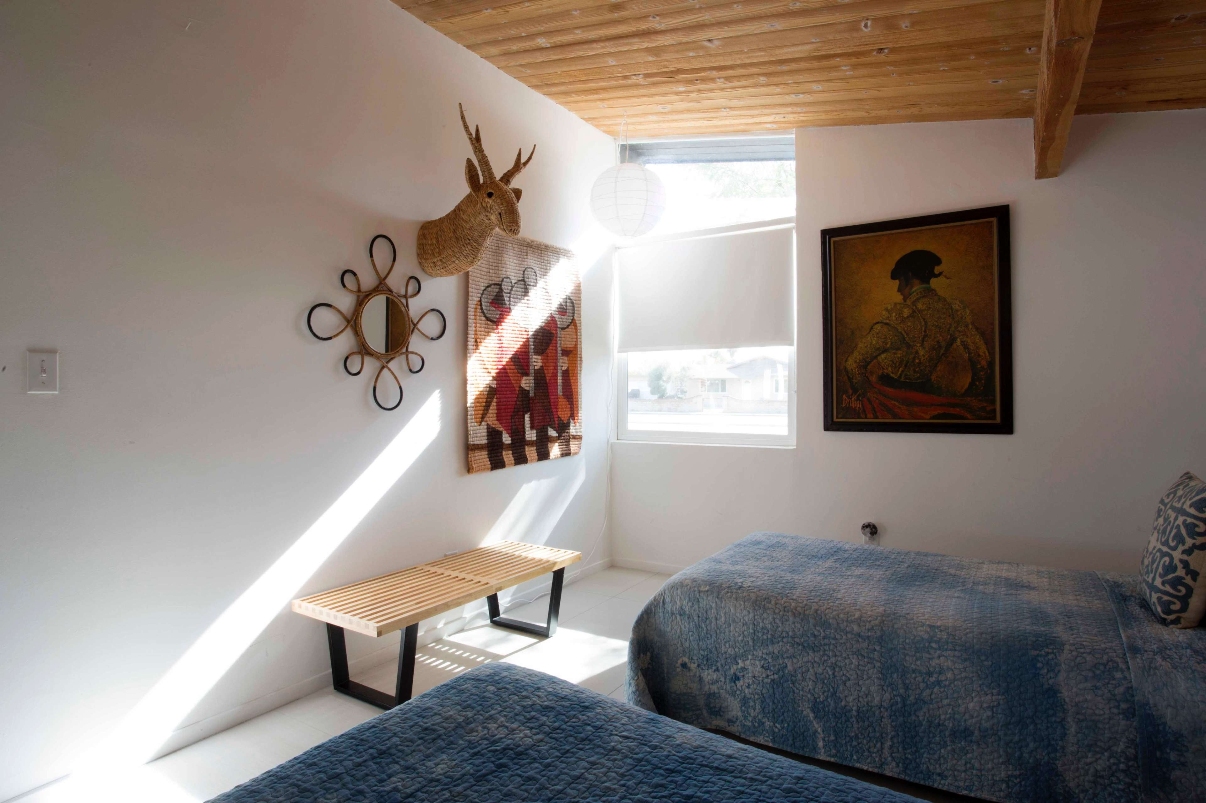 A bright bedroom with two beds, a decorative wall hanging, a mounted deer head, and a bench beside a window.