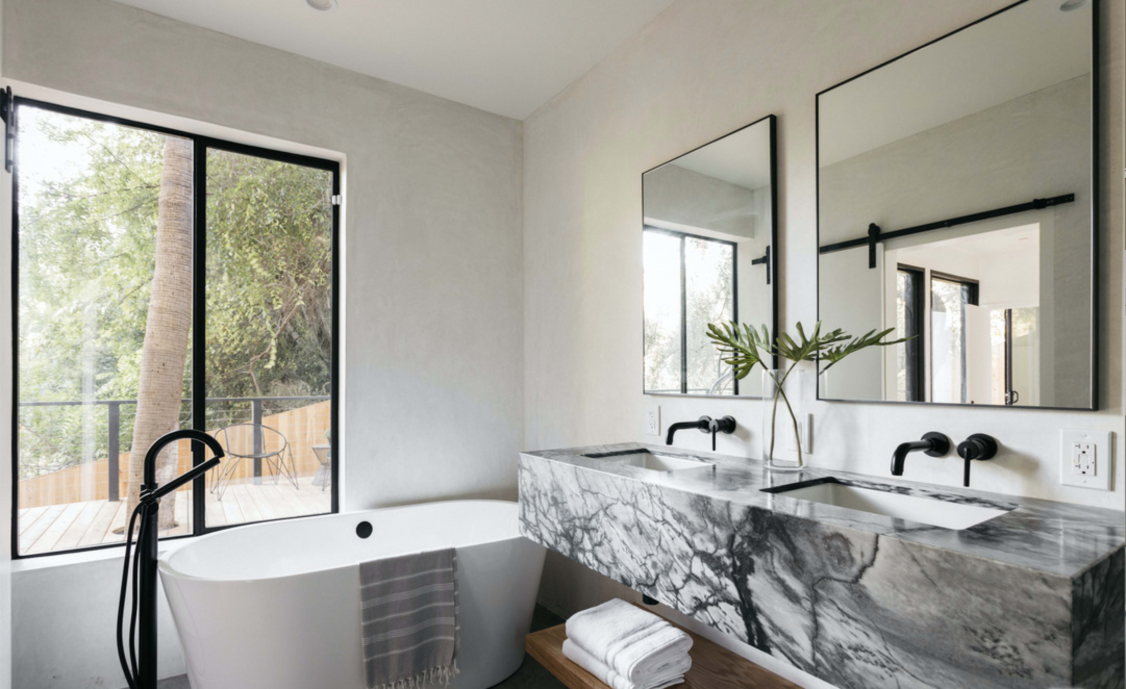 The bathroom features a freestanding tub, a double sink with a marble countertop, and large windows overlooking a wooden deck.