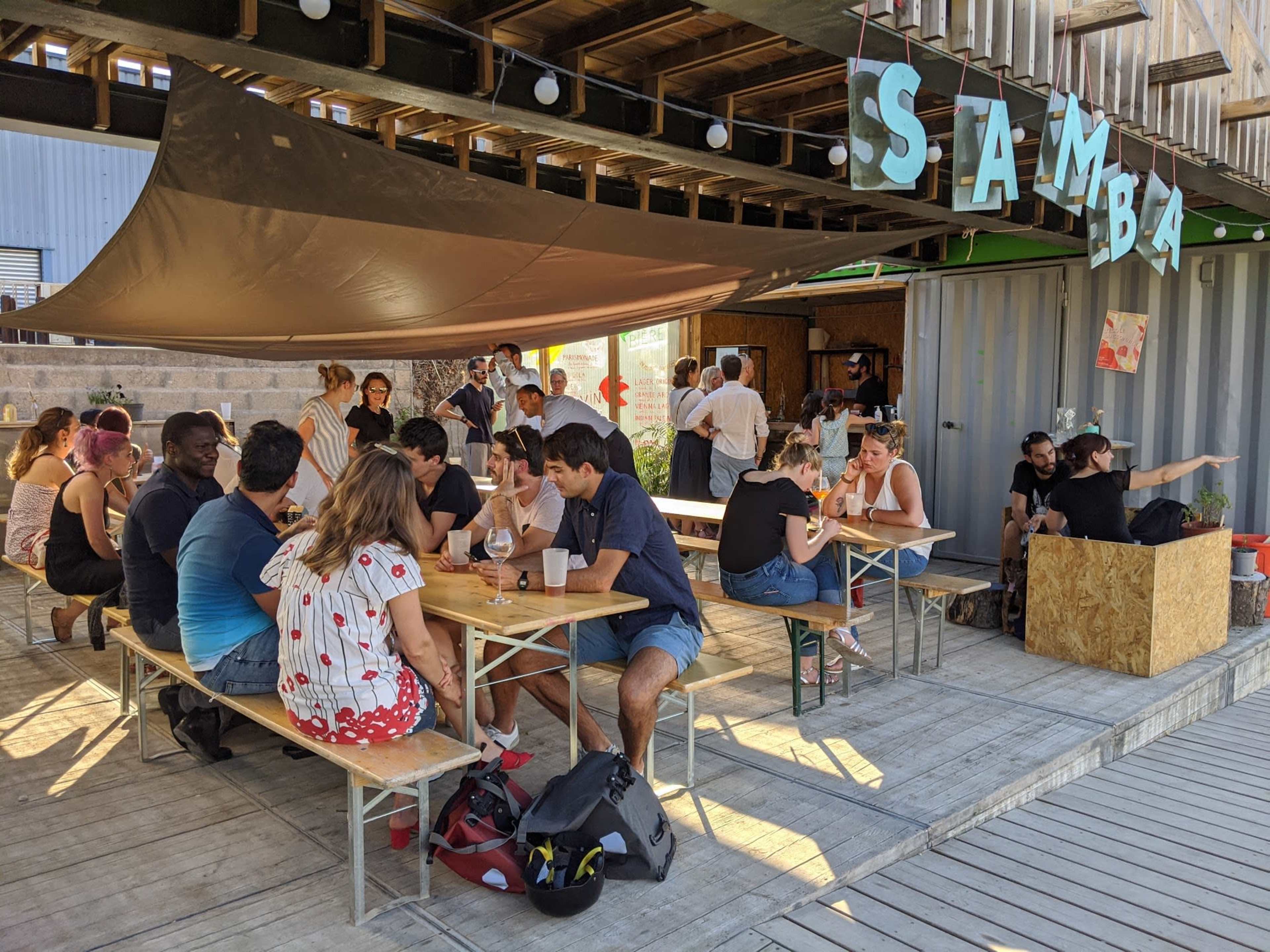 A group of people socialize around several tables under a canopy at an outdoor venue named "Samba."