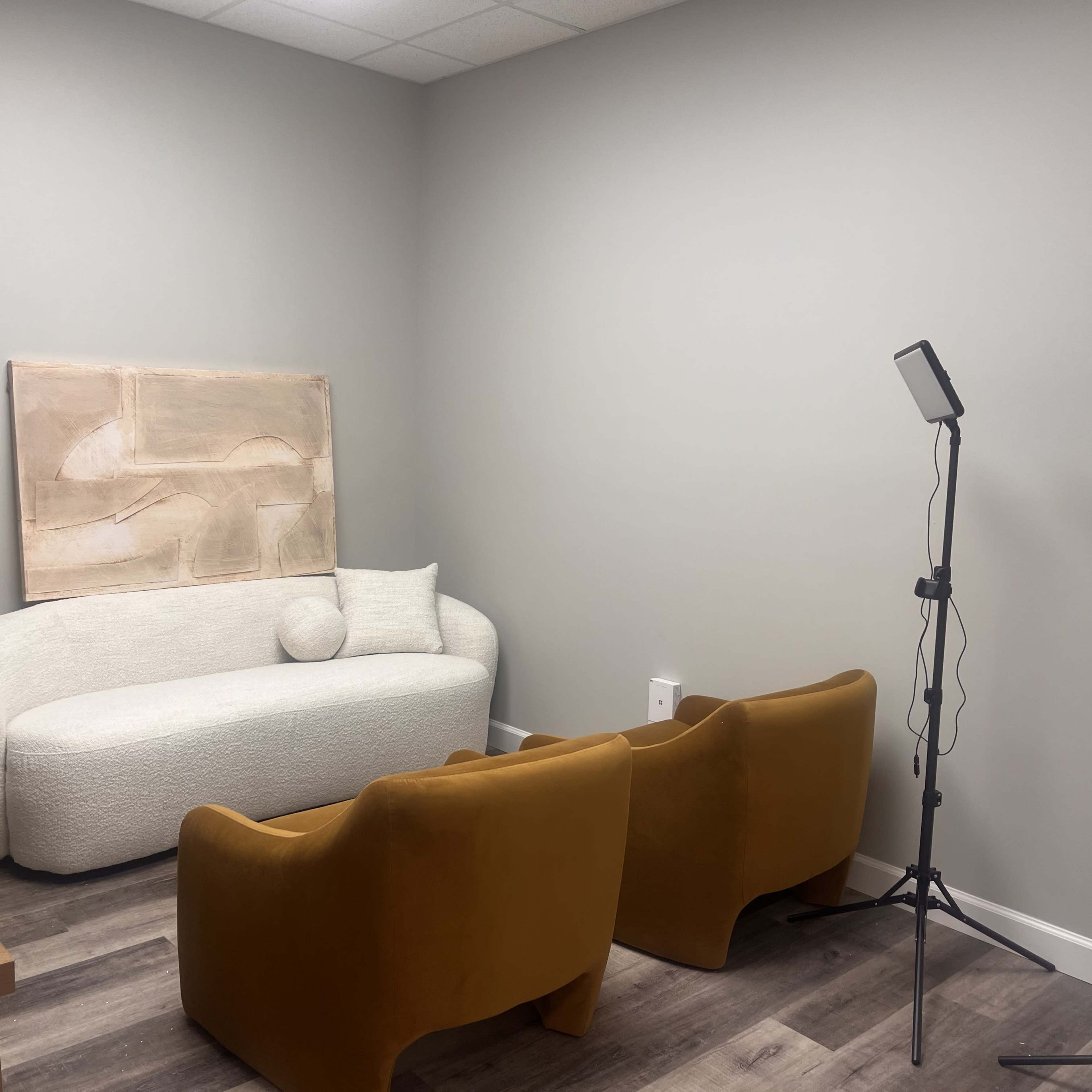 The image shows a small room with a curved white sofa, two orange chairs, and a floor lamp on a stand.