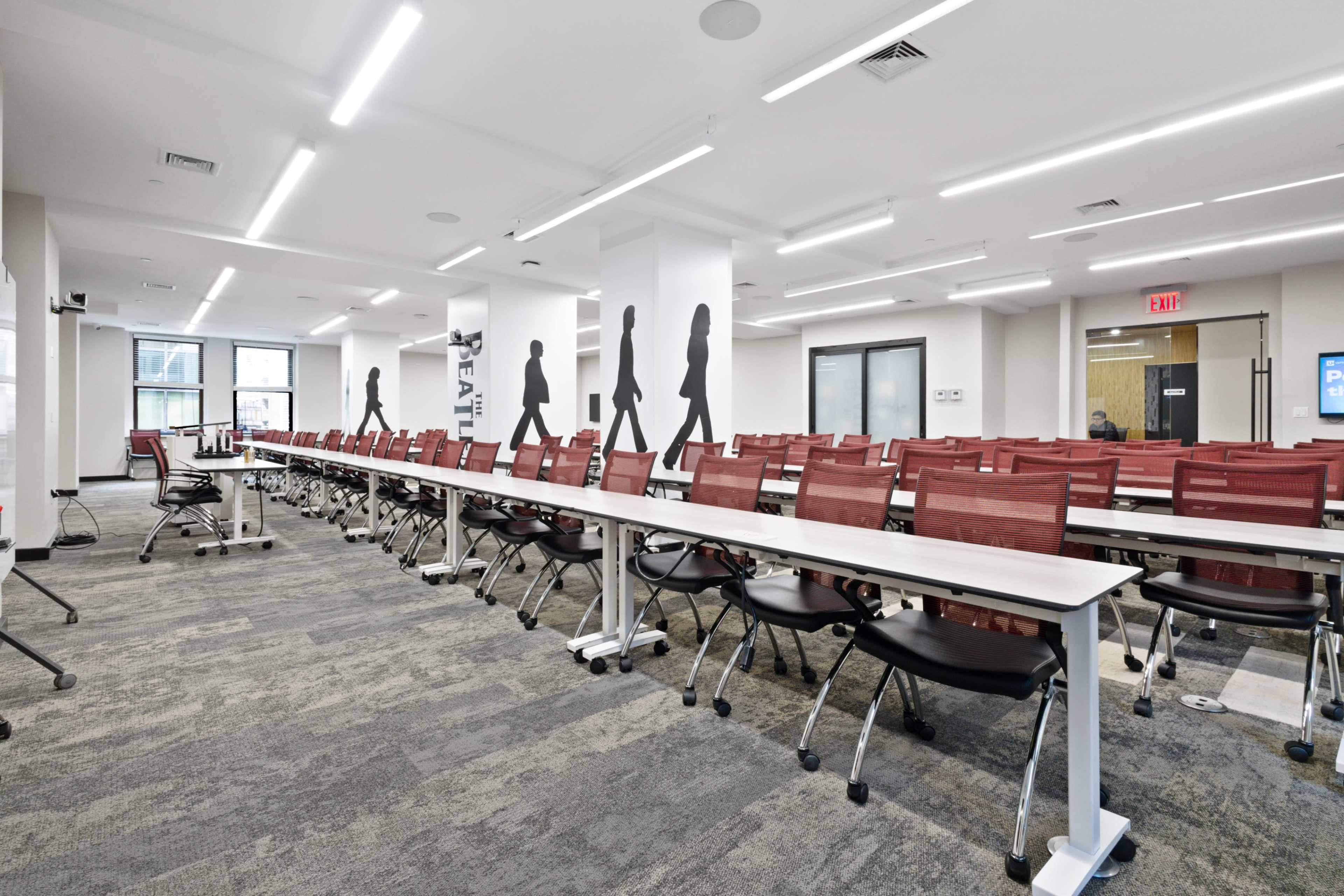 The image shows a modern classroom with rows of red chairs and white tables, featuring large windows and wall graphics of walking figures.