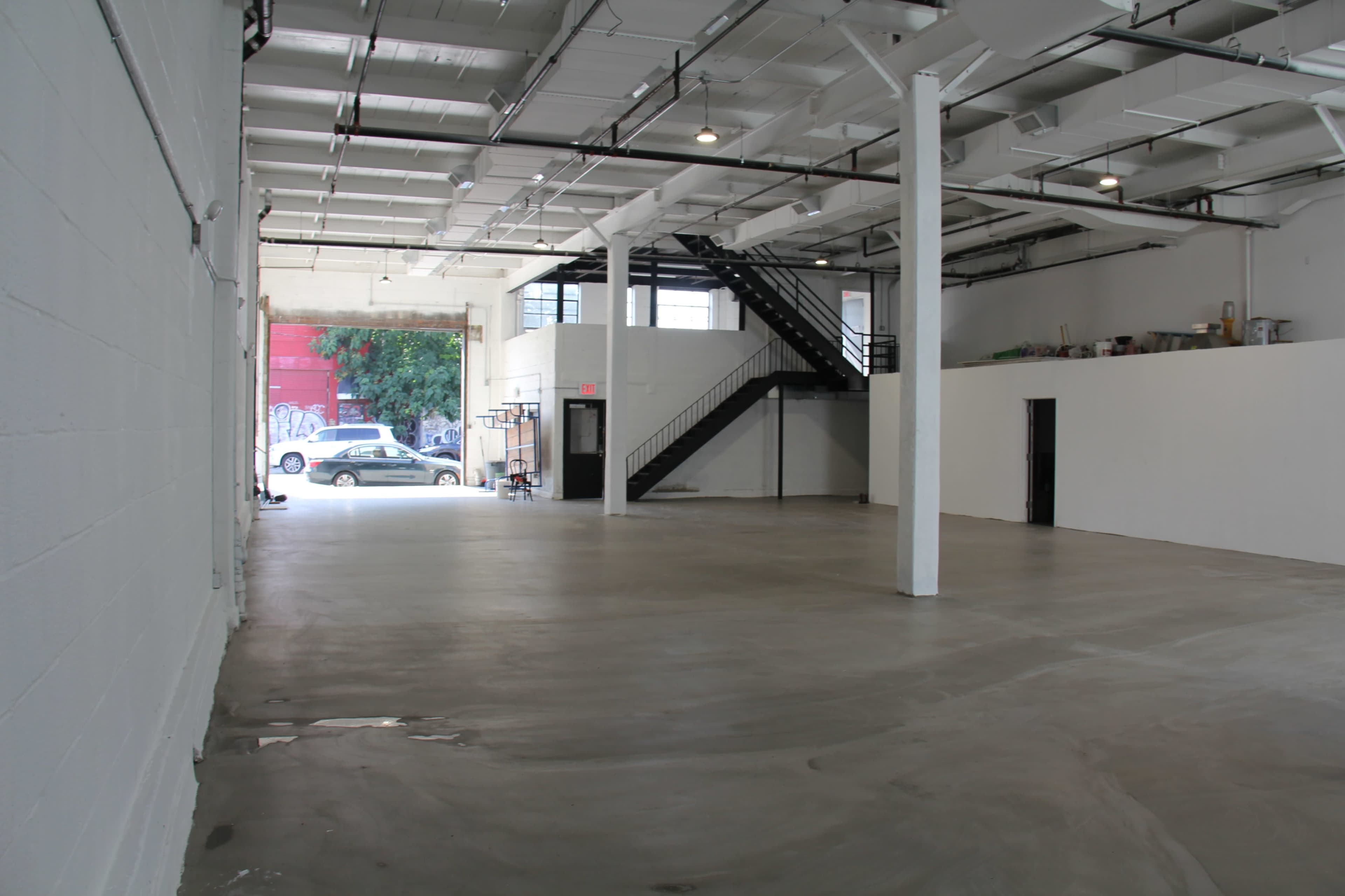 An empty warehouse space features a concrete floor, white walls, and a staircase in the background, with natural light coming from the entrance.