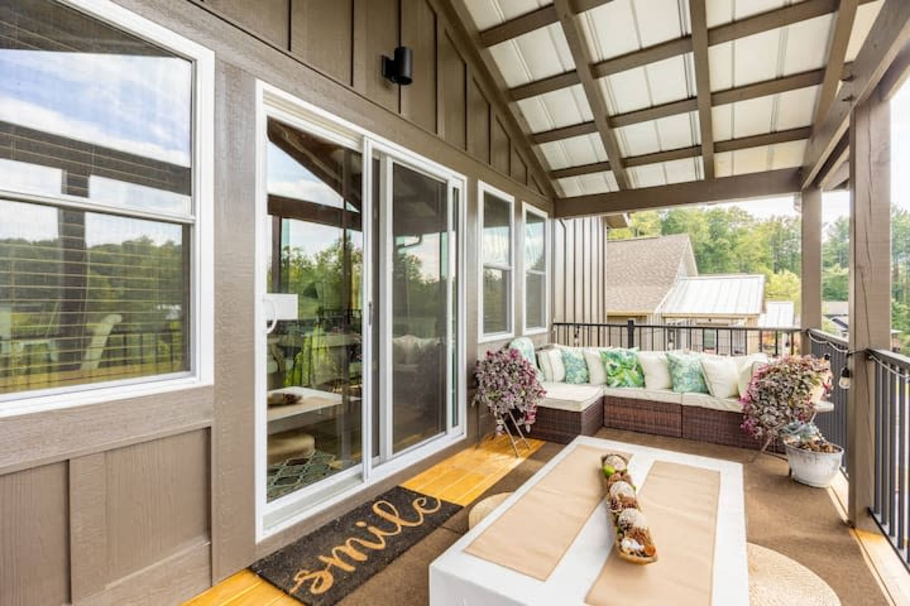 A porch area with a wicker sectional sofa, decorative cushions, and a coffee table, surrounded by green trees and a residential setting.