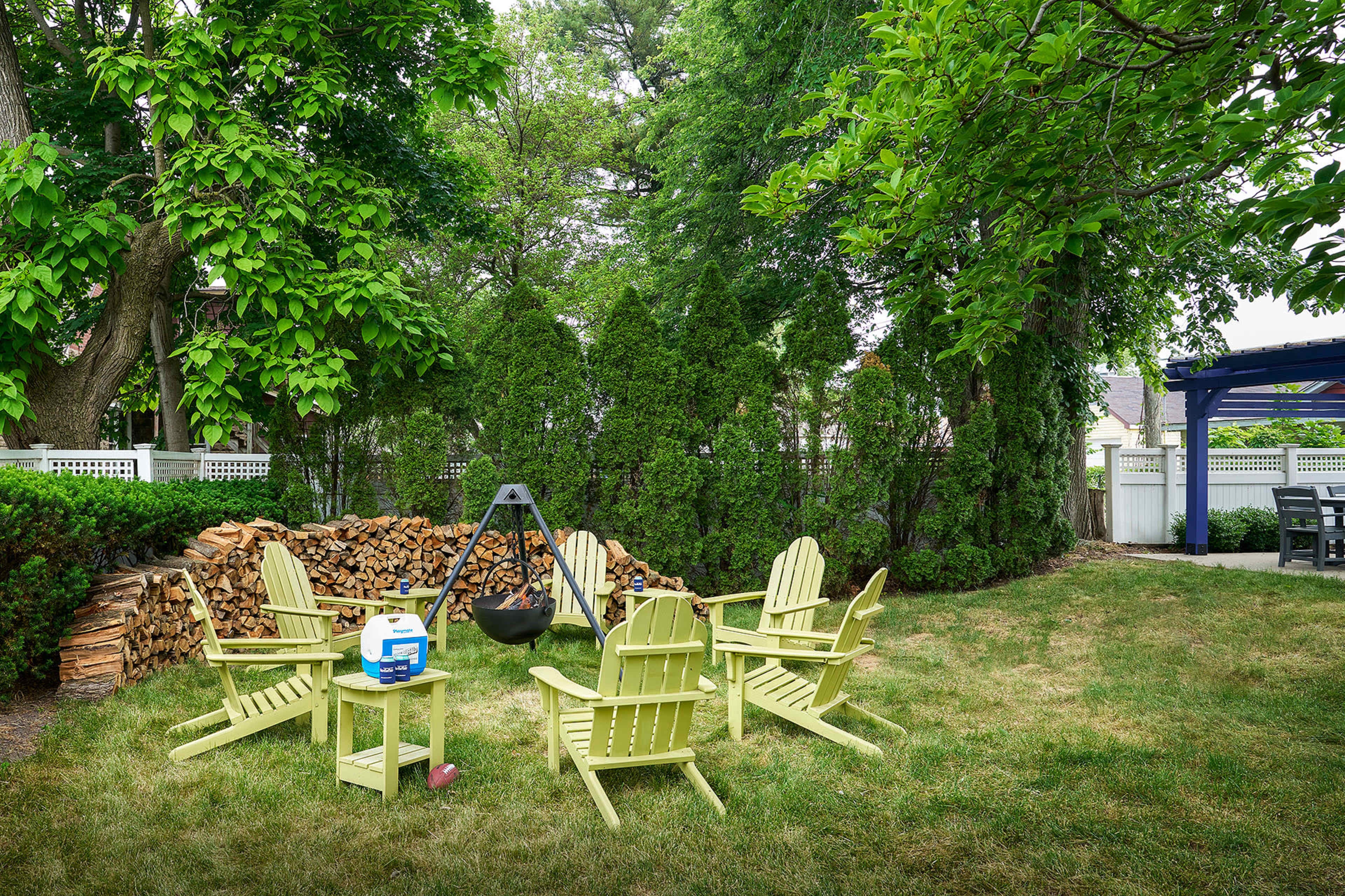 A circular arrangement of light green Adirondack chairs surrounds a fire pit in a grassy yard, with a stack of firewood and tall hedges in the background.