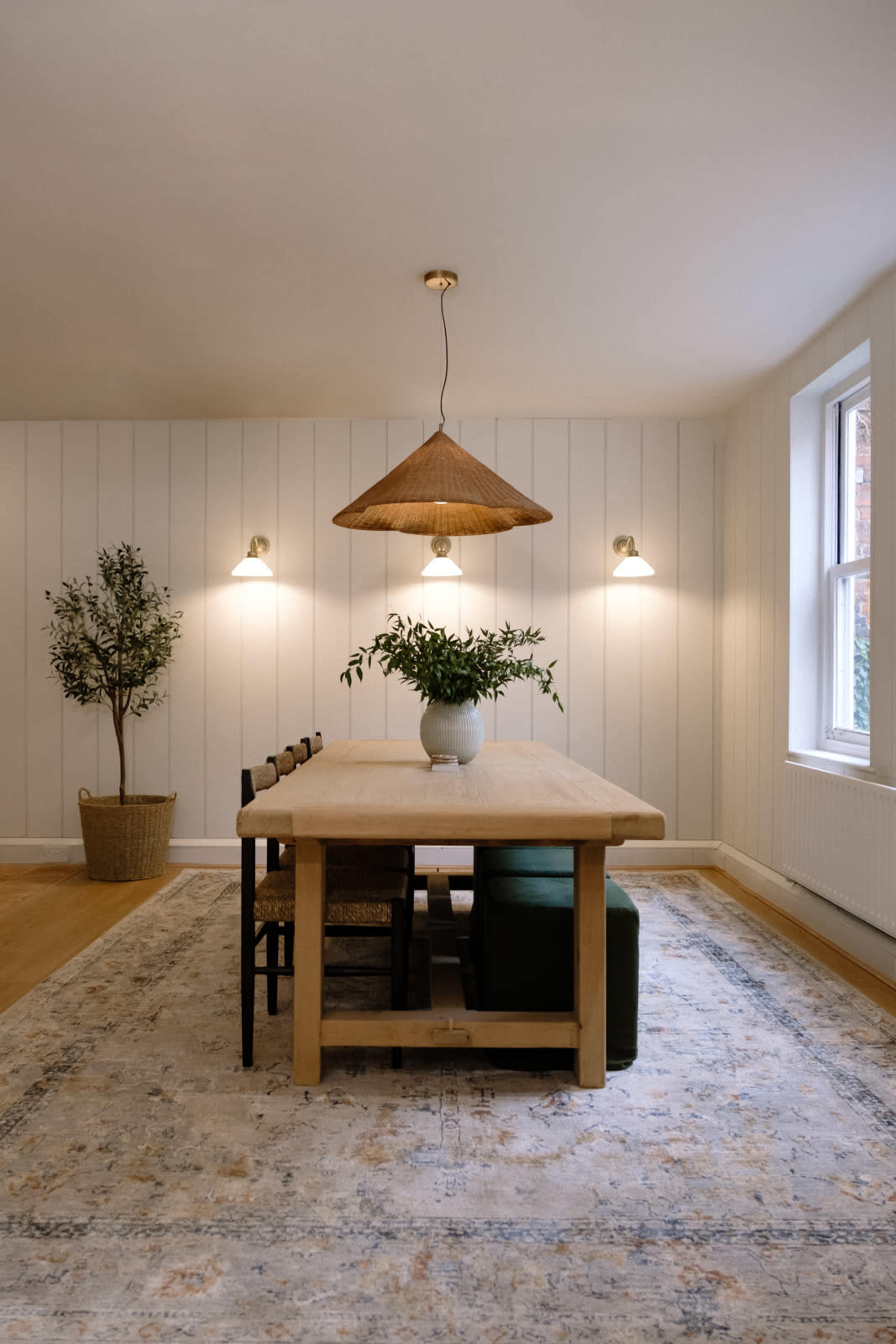 A wooden dining table is set in a softly lit room featuring white walls, green seating, and a hanging pendant light with a woven shade.