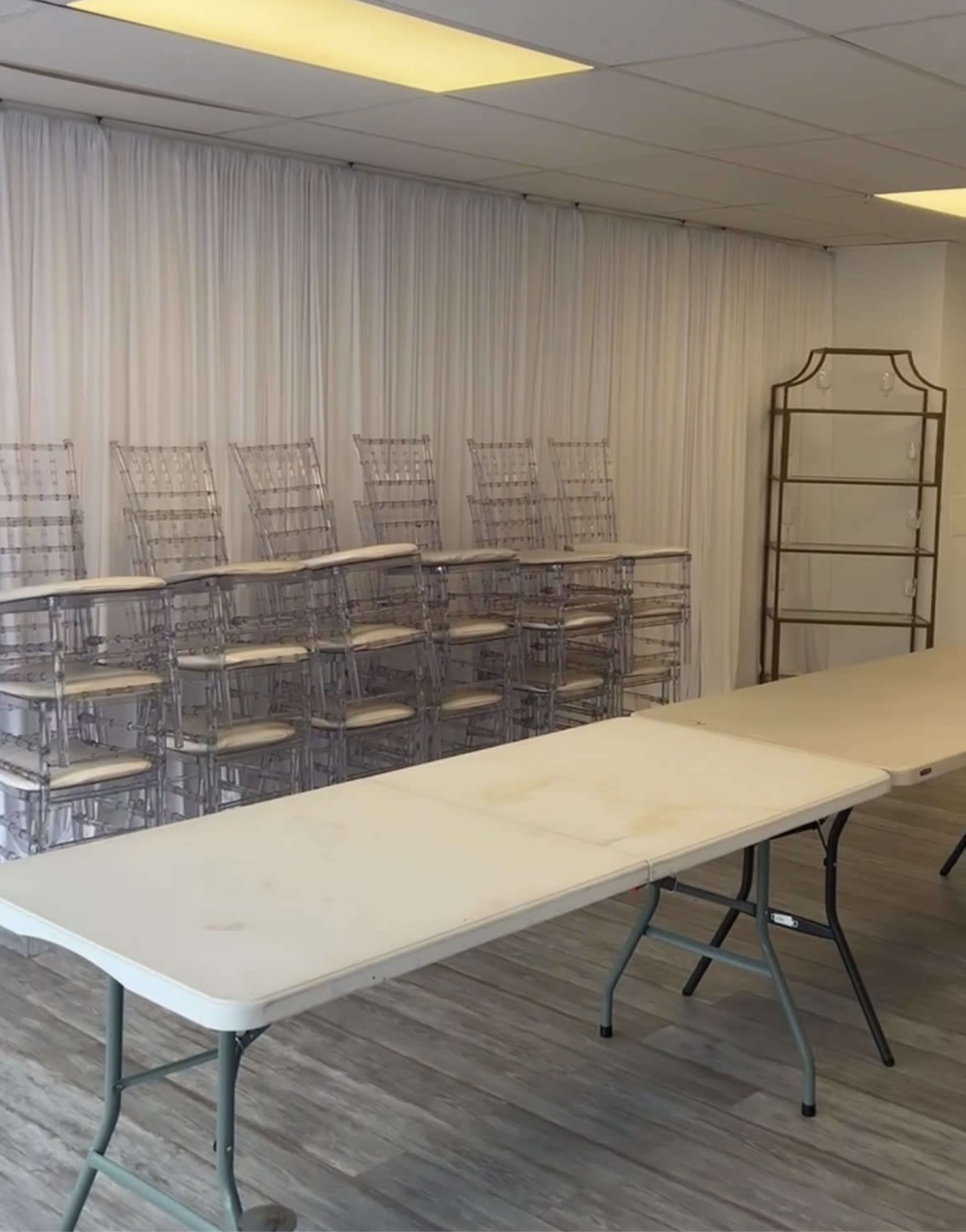 The image shows a room with several stacked transparent chairs against a fabric backdrop and two folding tables set up in front.