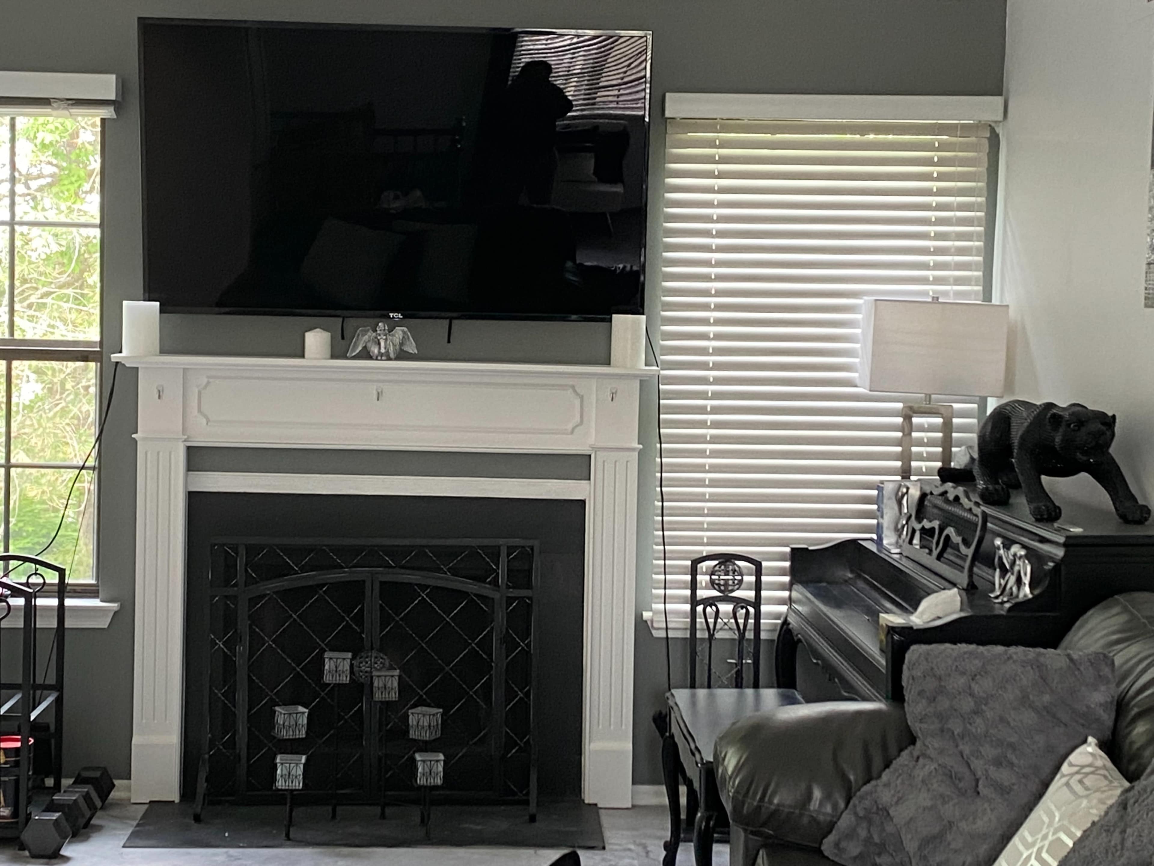A living room with a mounted television above a white fireplace, flanked by two windows with blinds, and a black piano beside a decorative black panther statue.