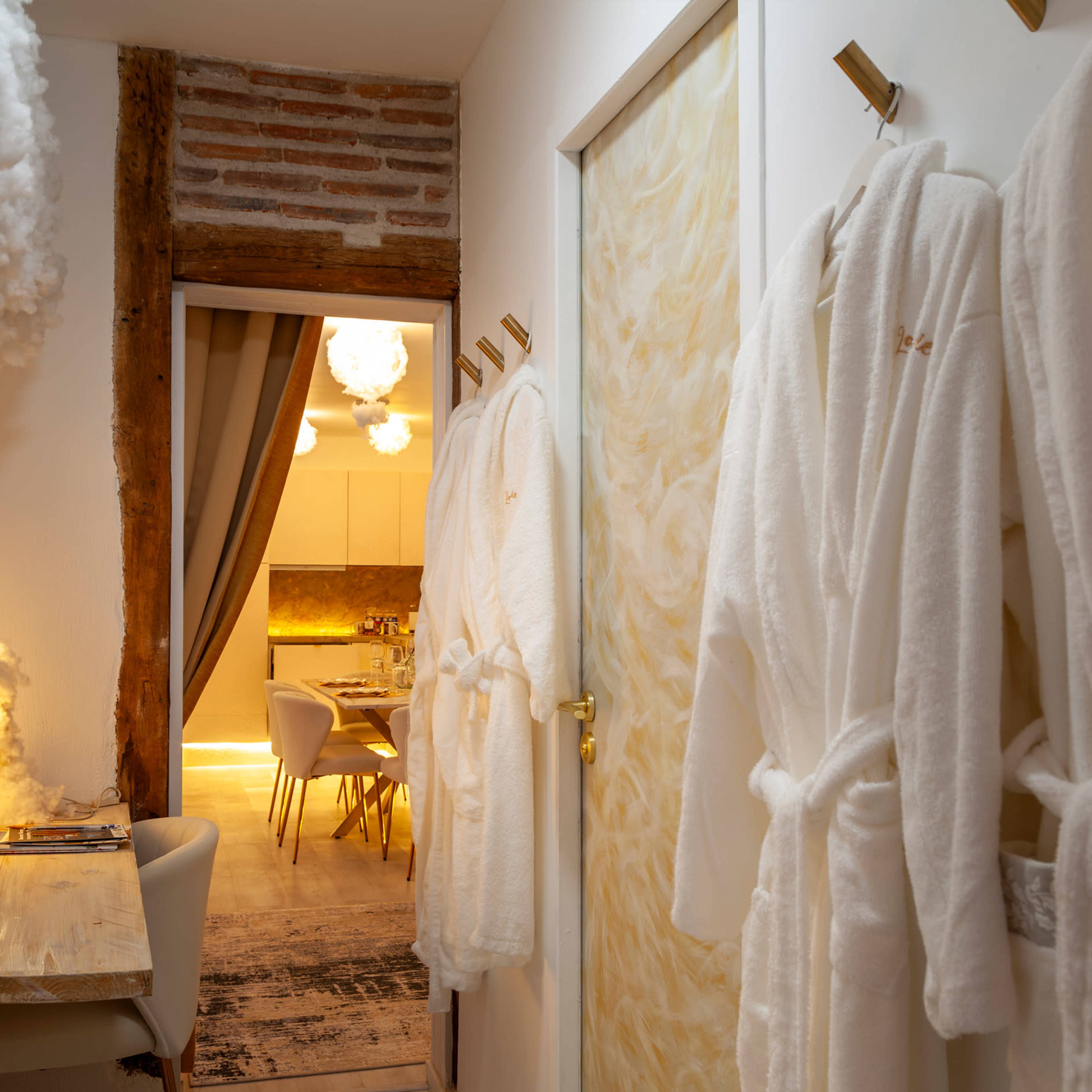 The image shows a narrow hallway with white robes hanging on hooks beside a door leading to a softly lit room with a dining area.