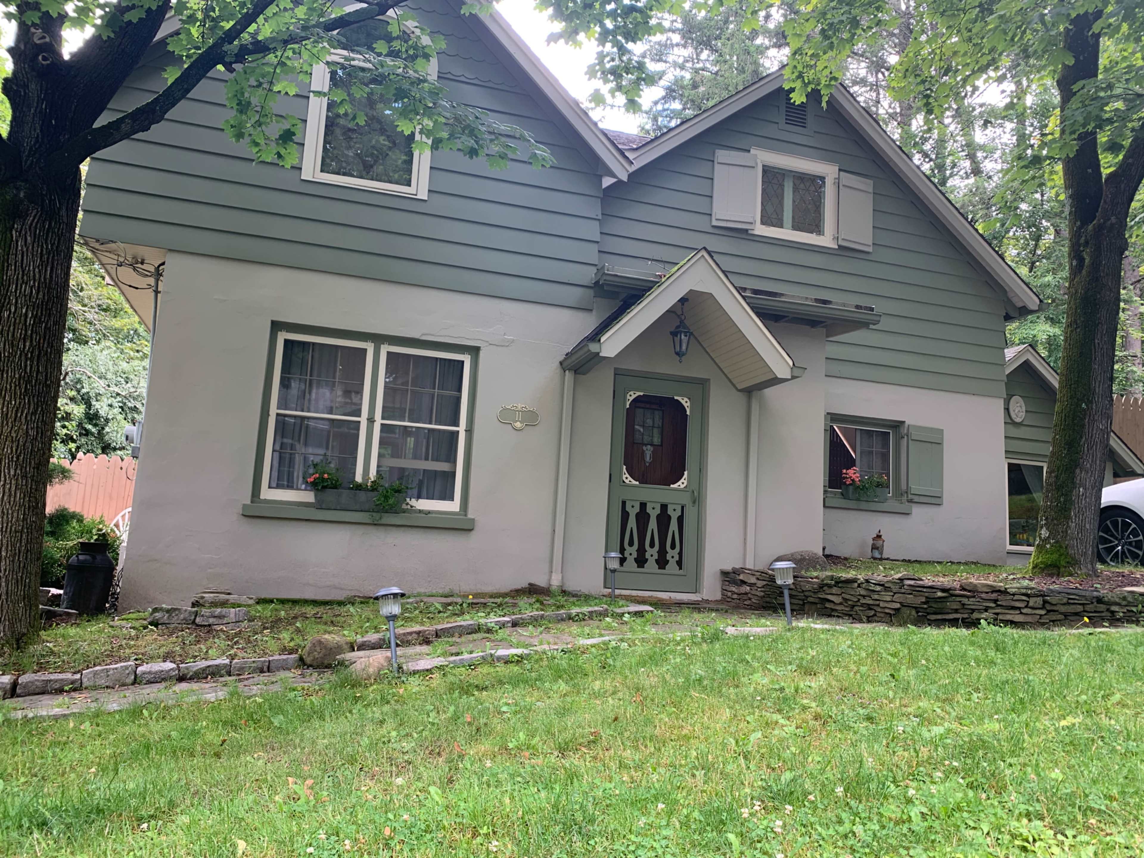 A two-story house with a green and white exterior, surrounded by grass and trees, and featuring a small stone pathway leading to the entrance.