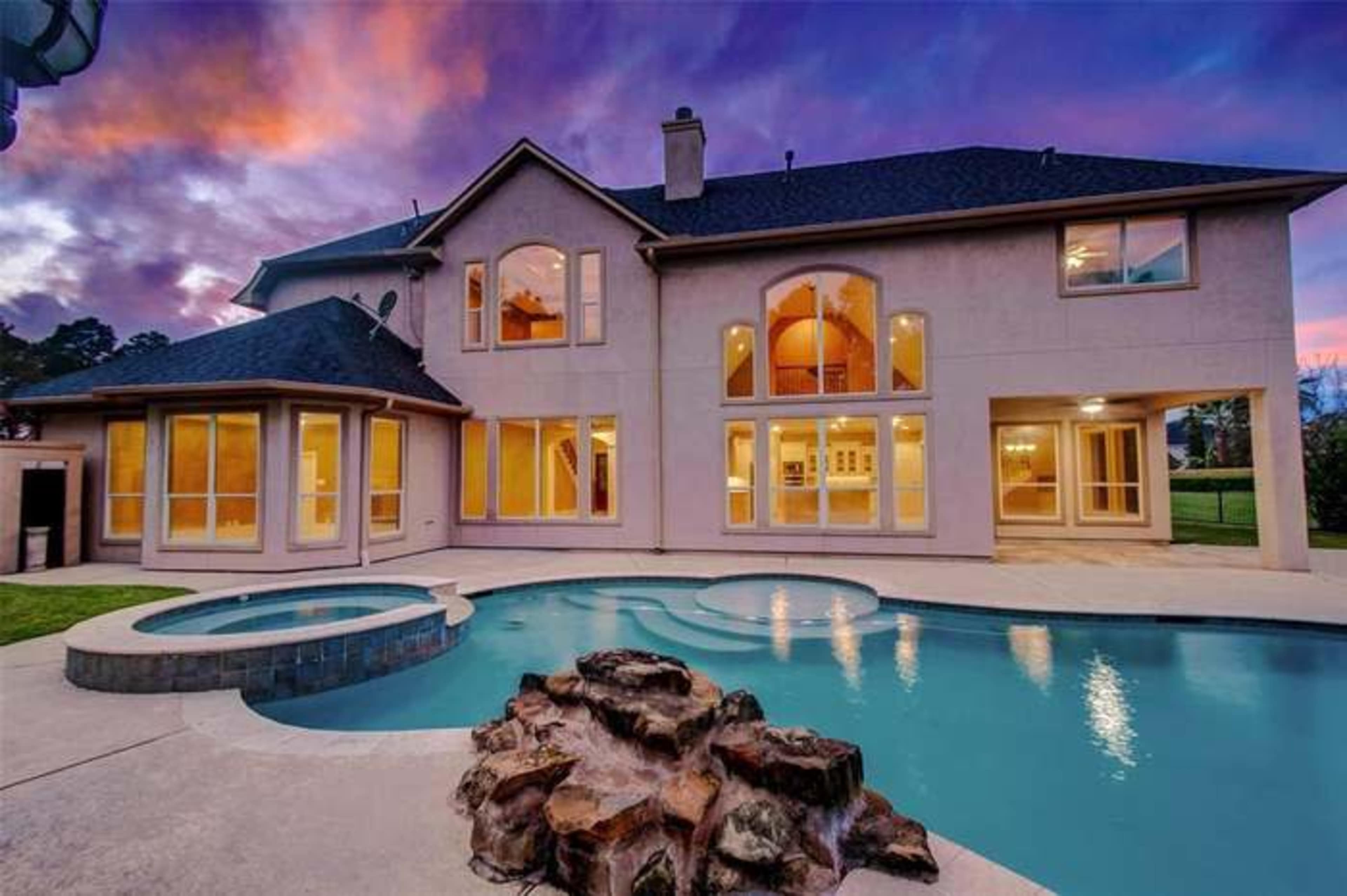 A modern two-story house with large windows, a swimming pool with a spa, and a landscaped backyard against a vibrant sunset sky.