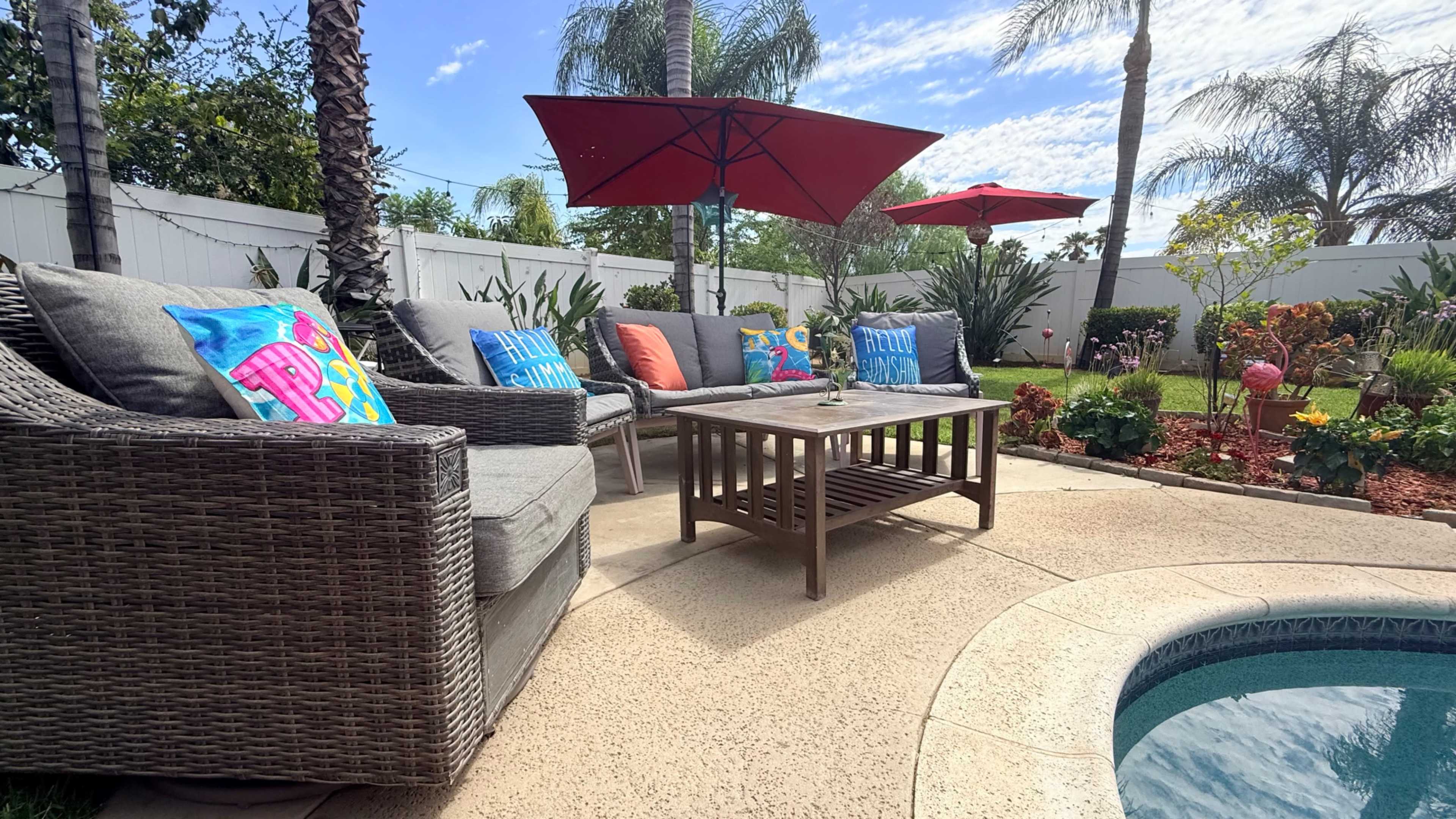 A backyard patio features a seating area with a table, colorful cushions, and two red umbrellas surrounded by lush greenery and a pool.