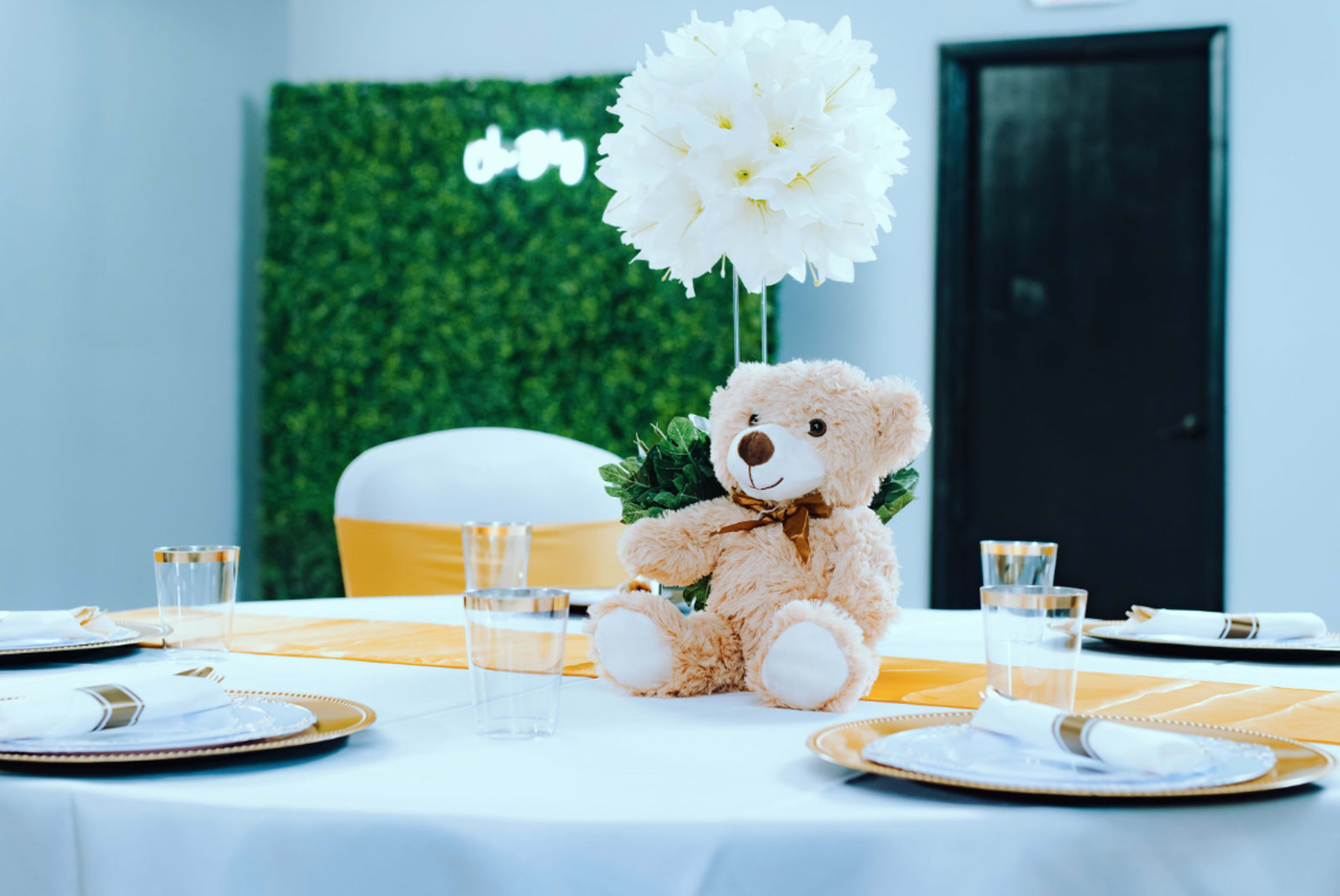 A round table is set with yellow and white decorations, featuring a teddy bear at the center and a large flower arrangement.