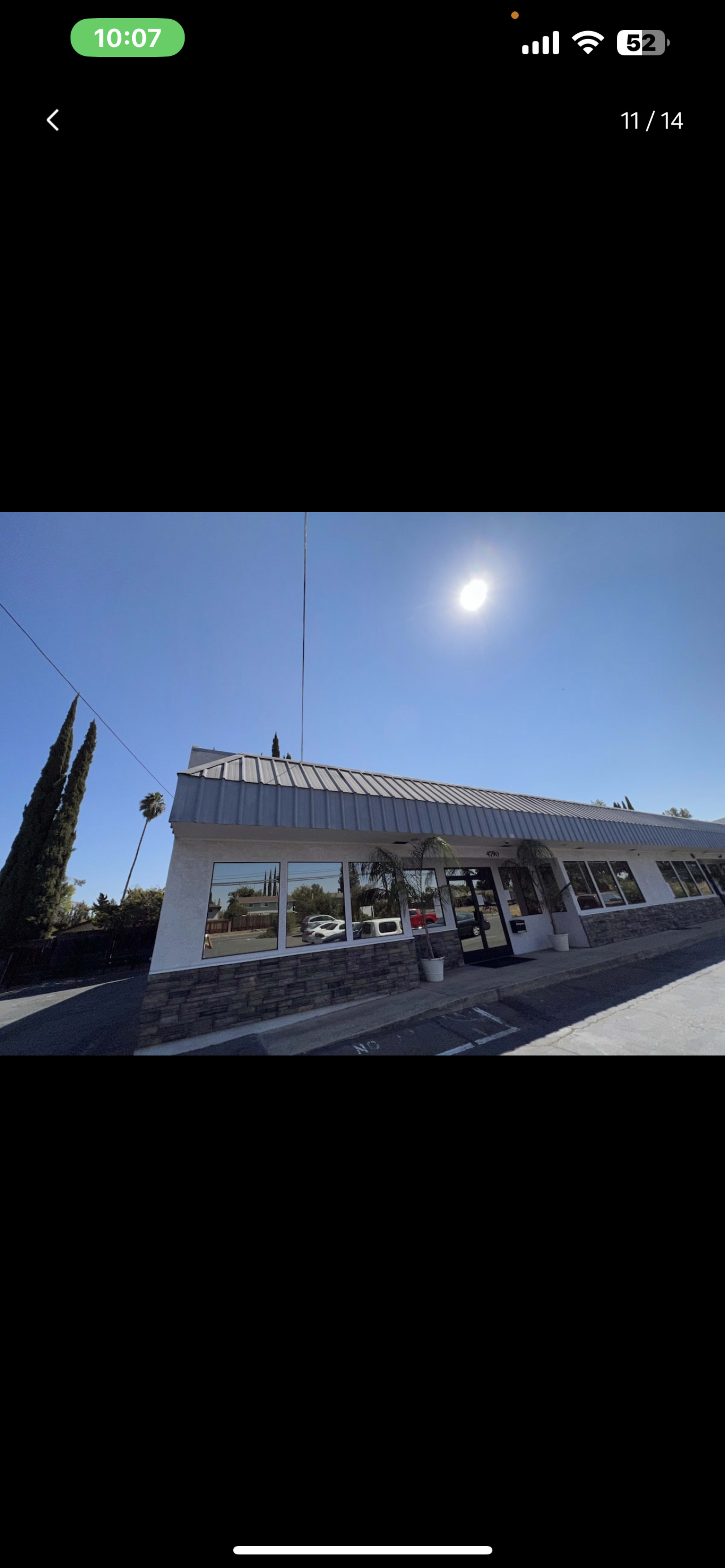 The image shows a single-story building with large windows, a peaked roof, and trees in the background under a clear sky.