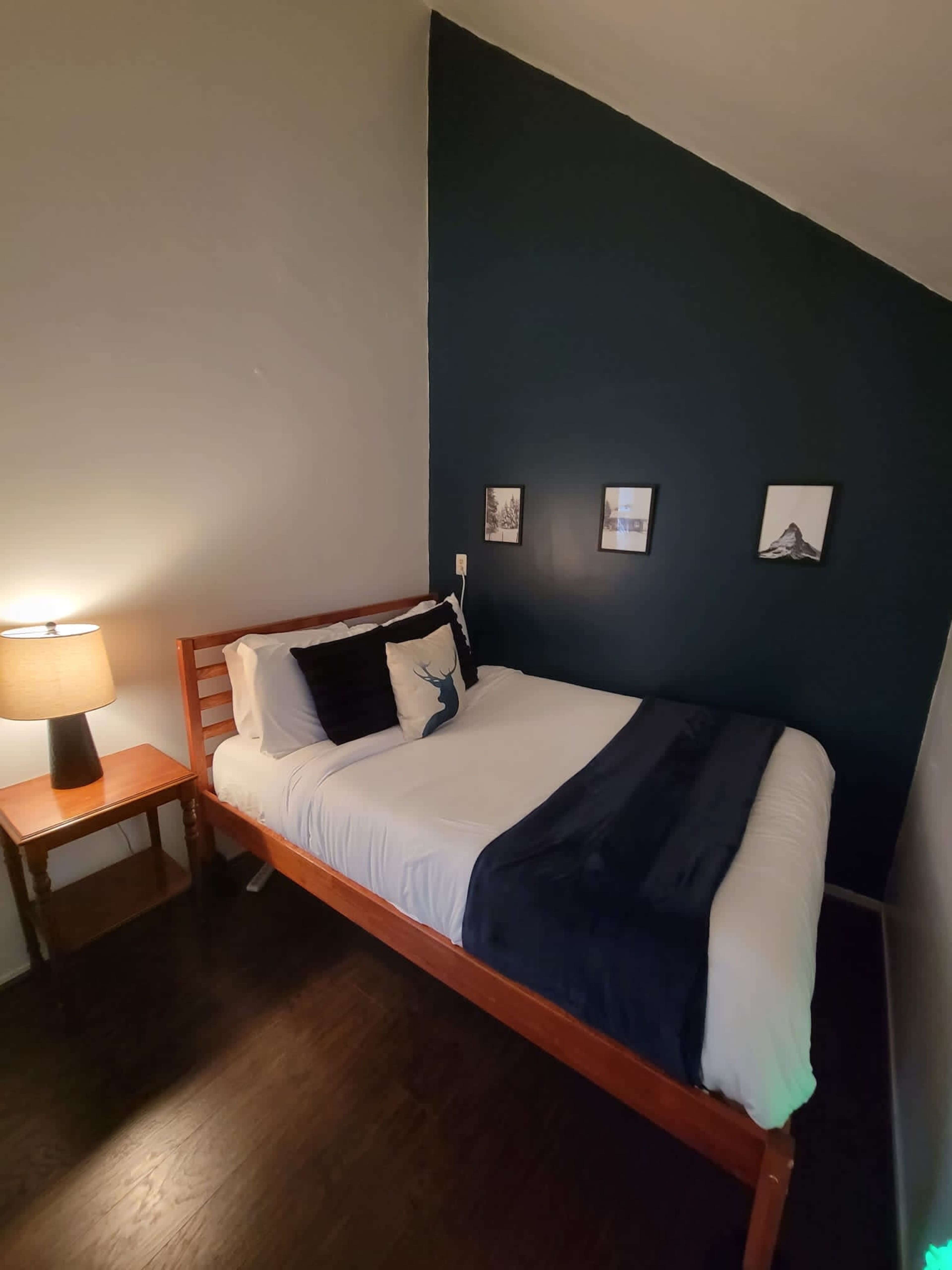 A small bedroom featuring a double bed with white bedding, a dark blue accent wall, a bedside table with a lamp, and framed pictures on the wall.