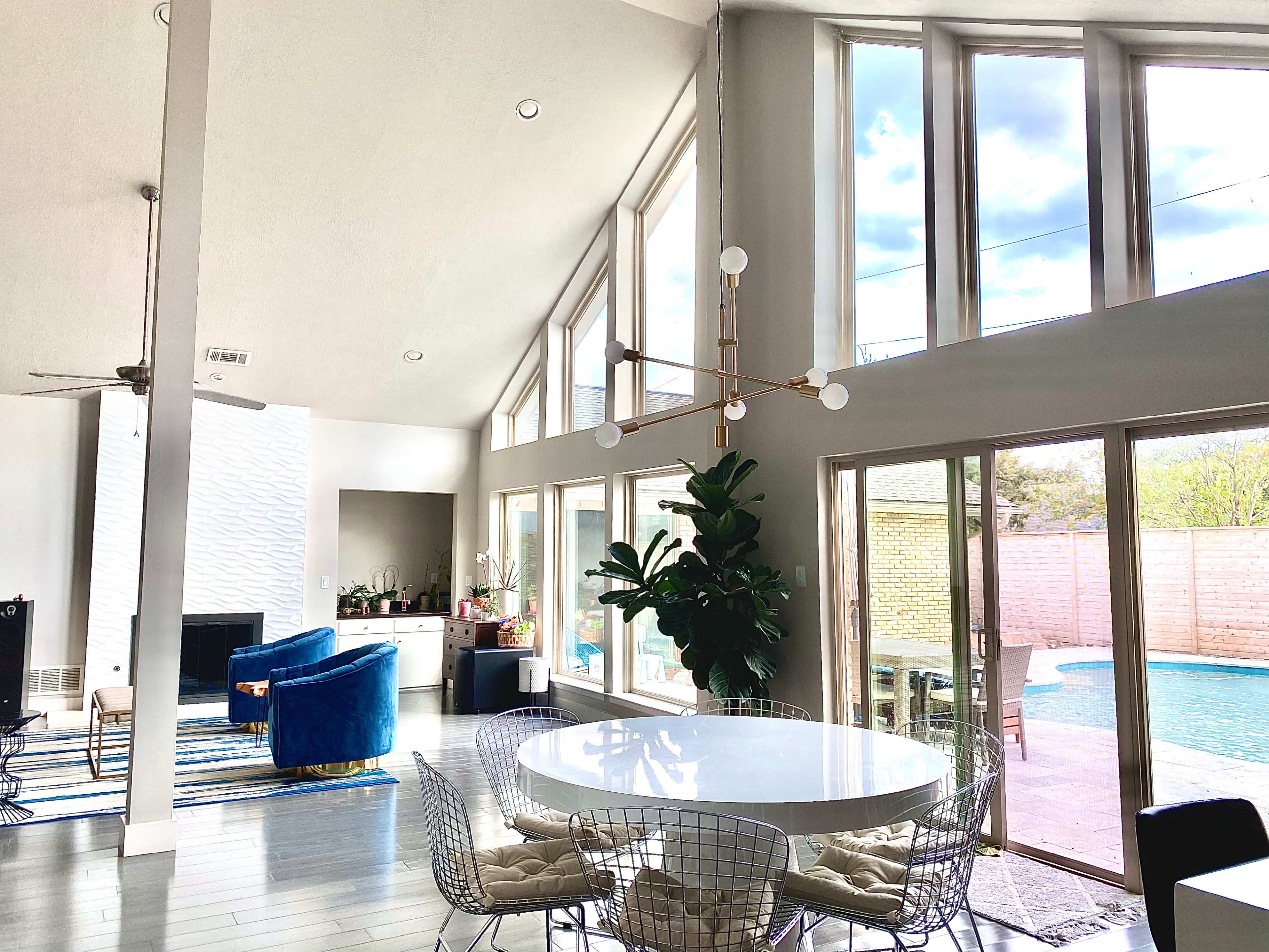 A modern living space with large windows, a circular dining table surrounded by wire chairs, and a view of a pool outside.