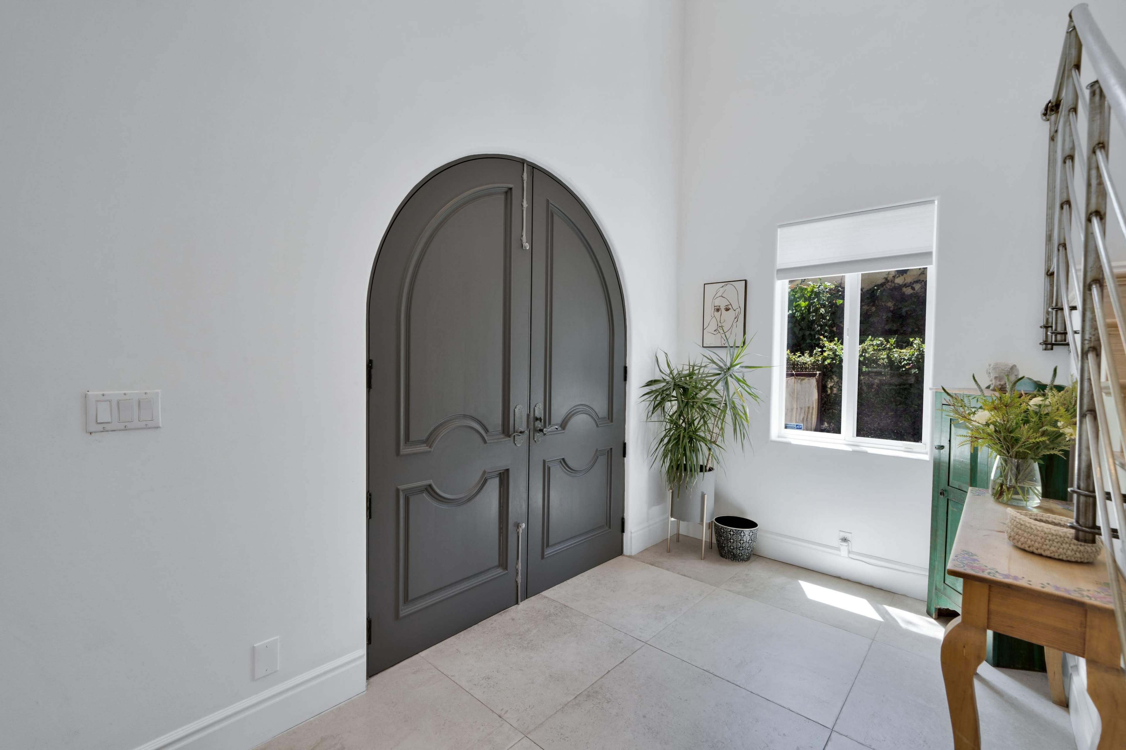 The image shows a bright entryway featuring large gray double doors, a window with a view of greenery, and a small table decorated with a vase of flowers.