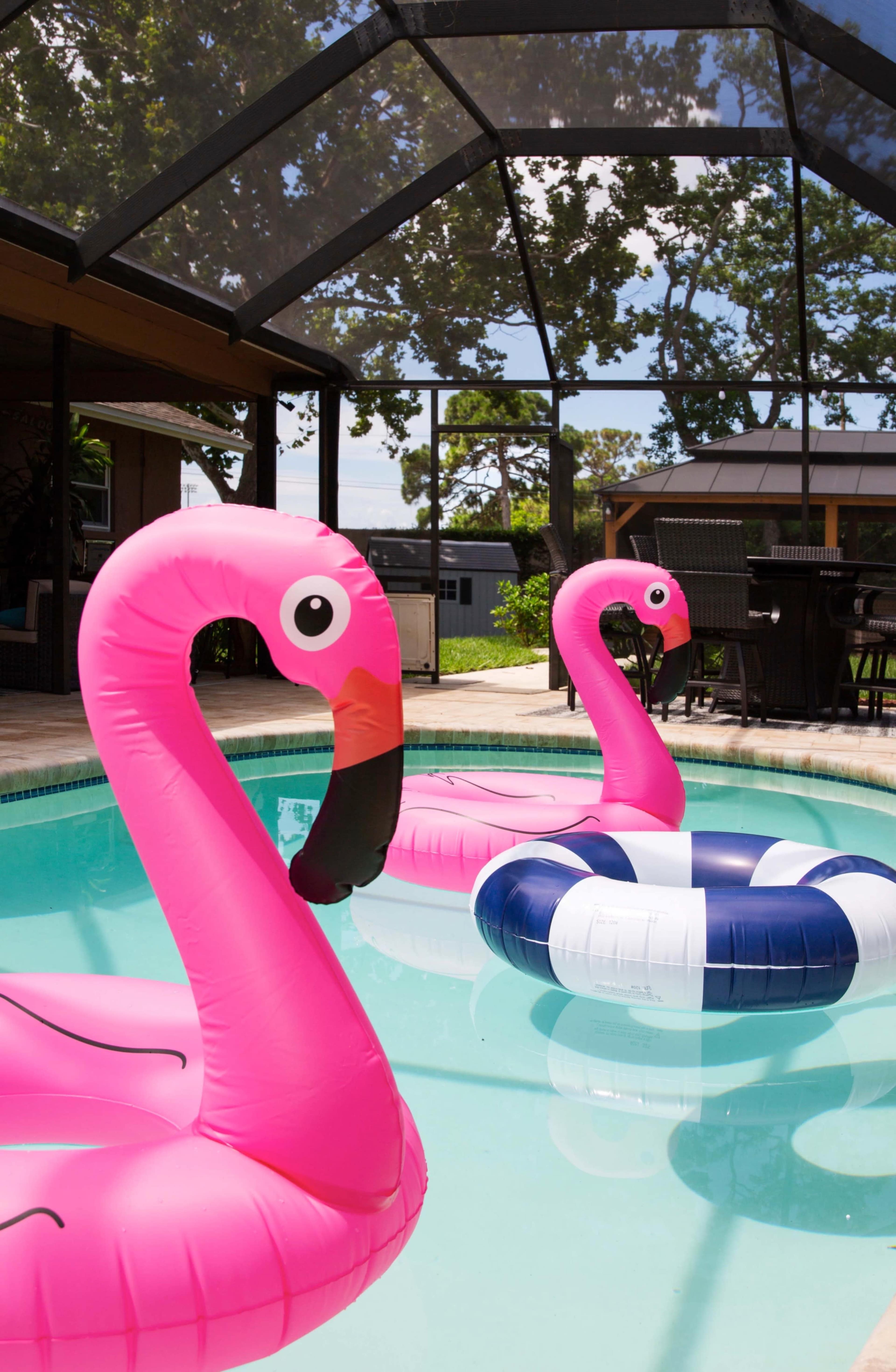 Two pink flamingo pool floats are partially submerged in a clear swimming pool surrounded by a screened patio.