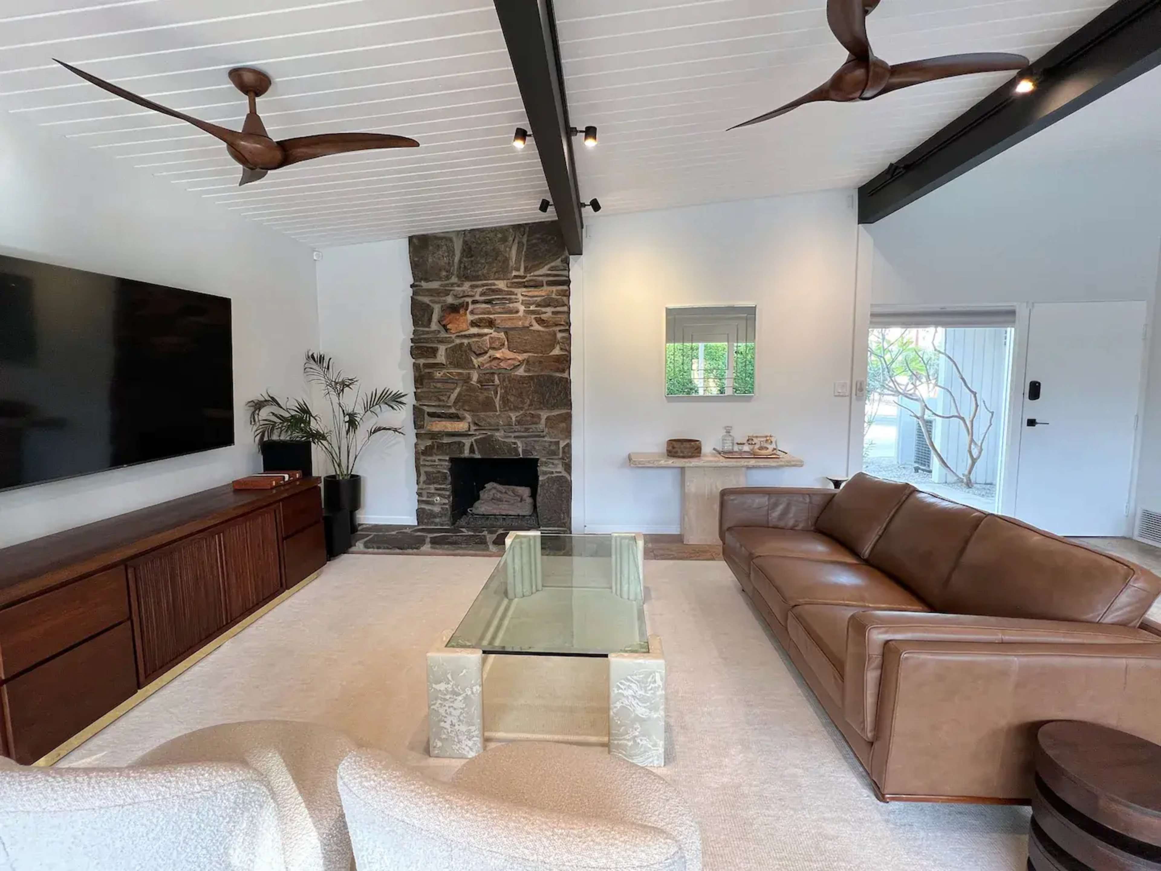 The living room features a stone fireplace, a large television mounted on the wall, and a comfortable leather sofa arranged around a glass coffee table.