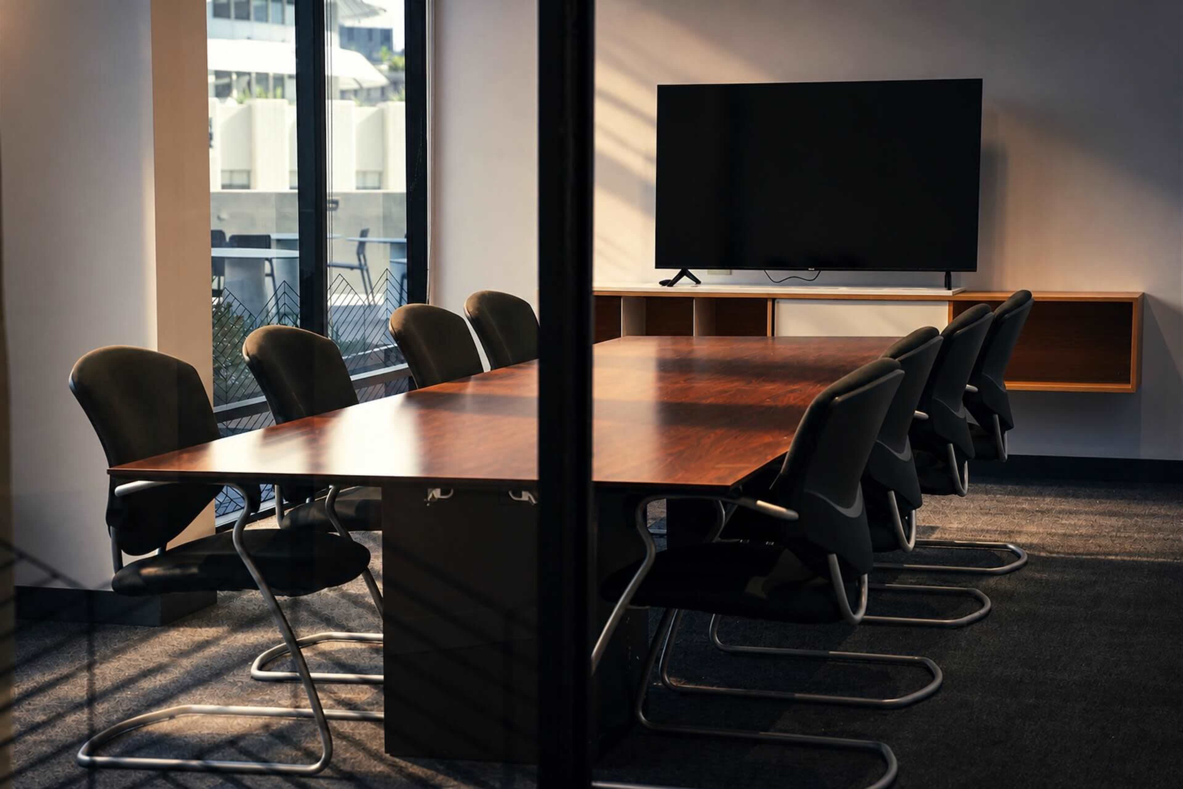 A modern conference room features a long wooden table surrounded by black office chairs, with a large flat-screen TV mounted on the wall.