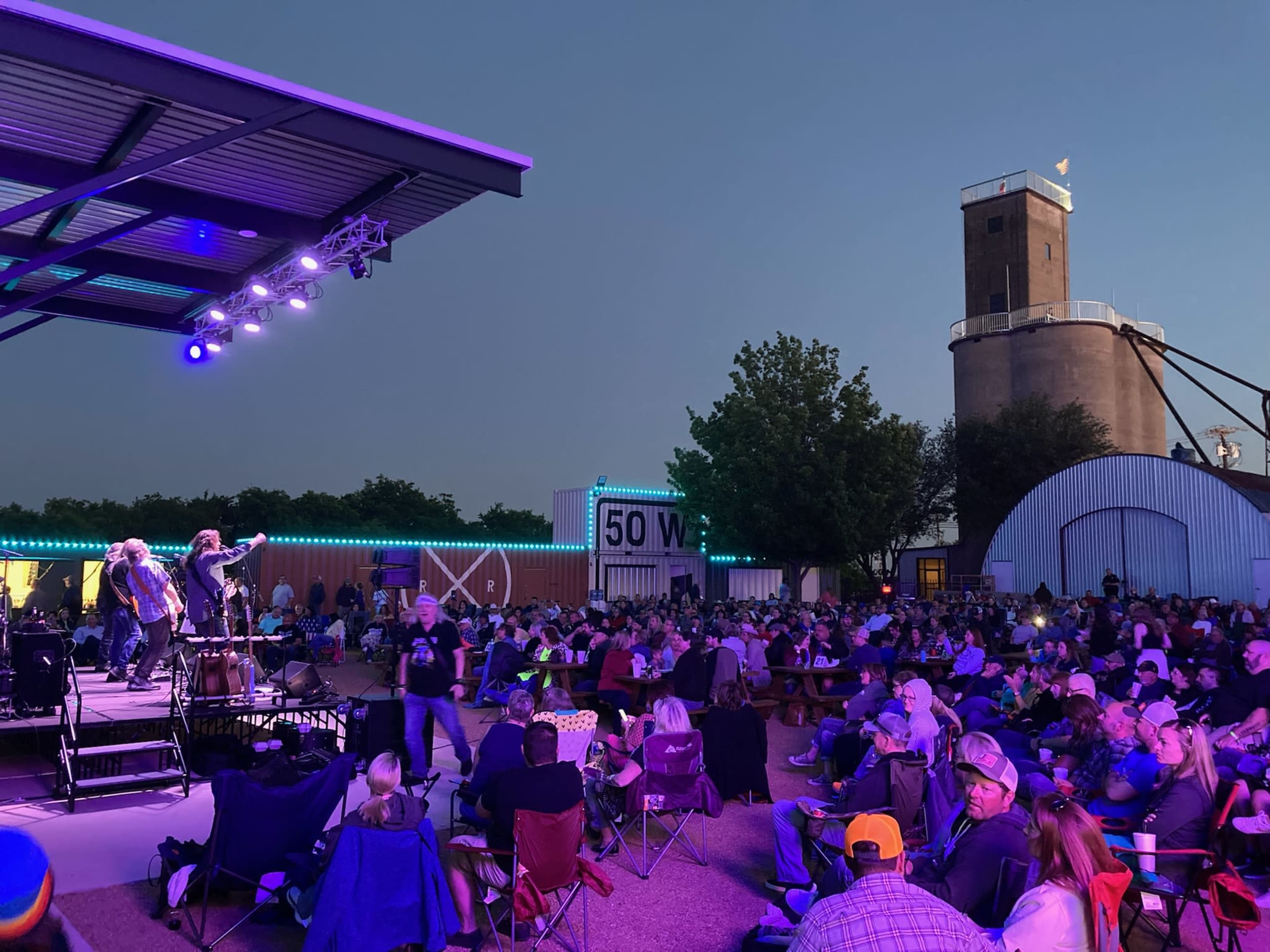 A large crowd gathers in an outdoor venue with performers on stage, illuminated by stage lights, while a historic tower looms in the background.