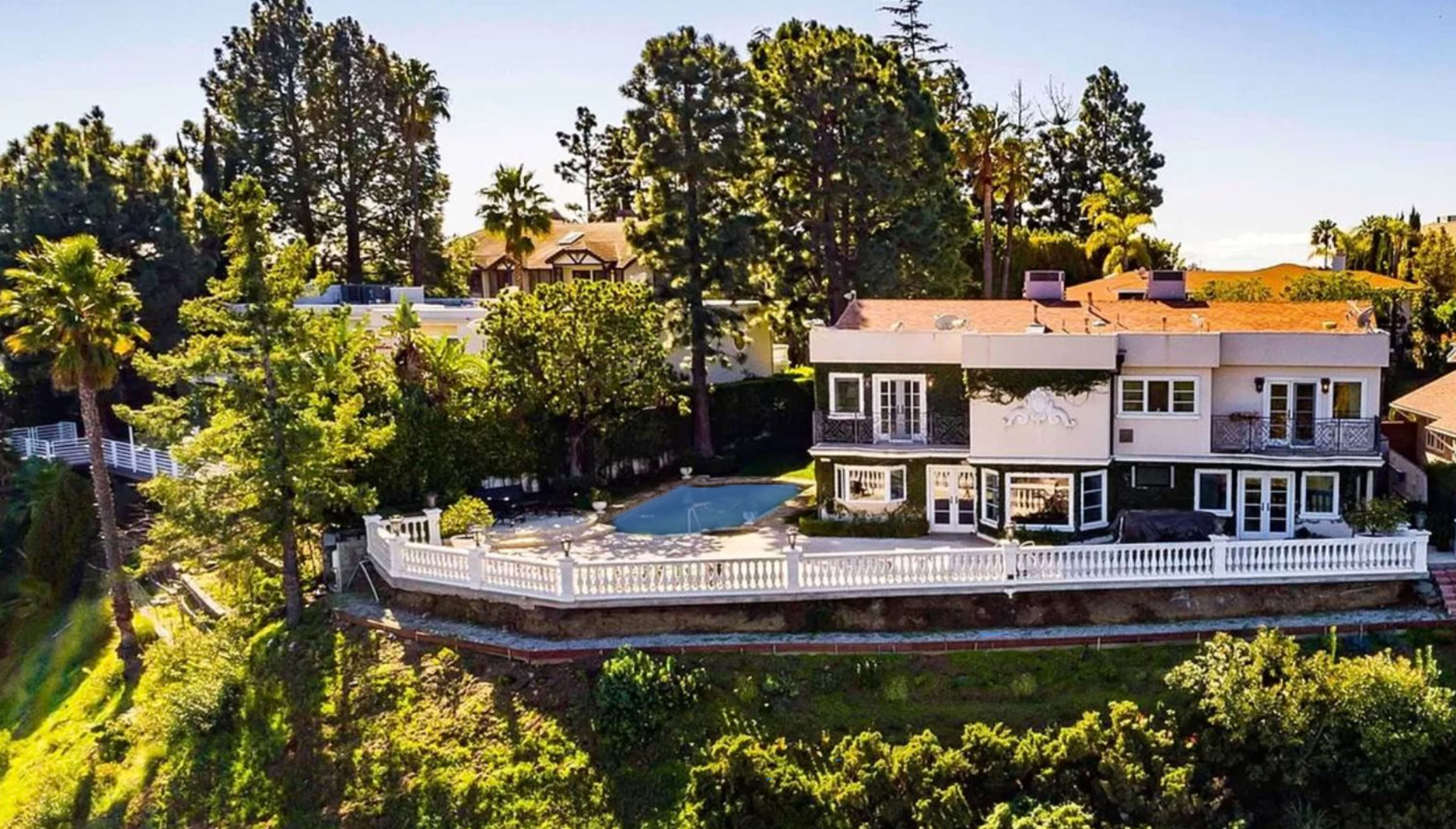 The image shows a large house situated on a hillside surrounded by trees and featuring a swimming pool in the yard.