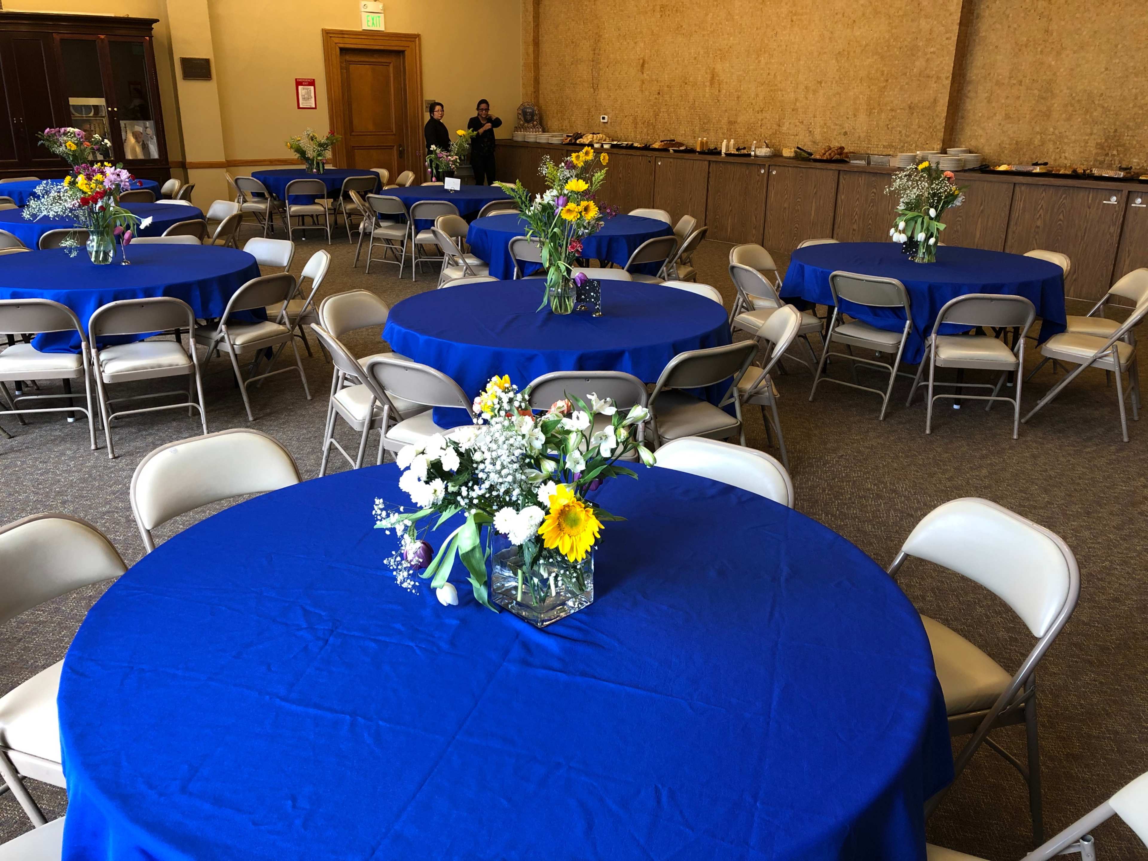 A dining hall features blue tablecloths draped over round tables, each adorned with floral centerpieces, while a buffet area is visible in the background.