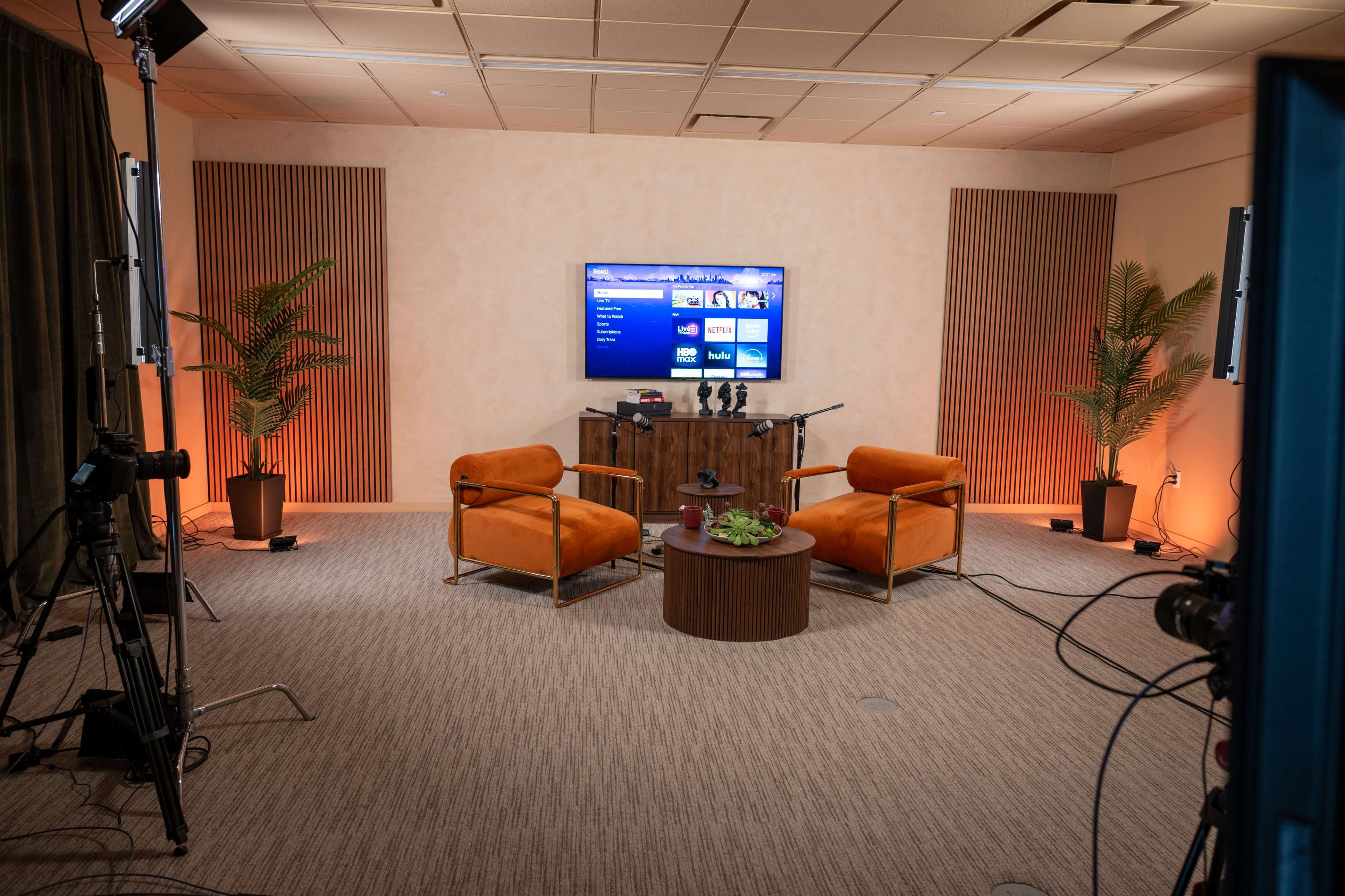 The image shows a modern room setup for a video recording, featuring two orange chairs, a small round table, and a television displaying content in a well-lit environment with plants in the corners.